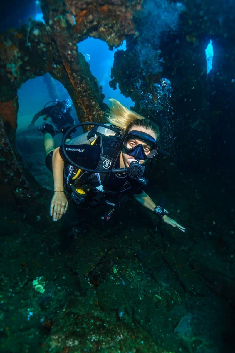 Diver with blonde hair and full scuba gear swimming through an underwater wreck structure as a second diver looks on