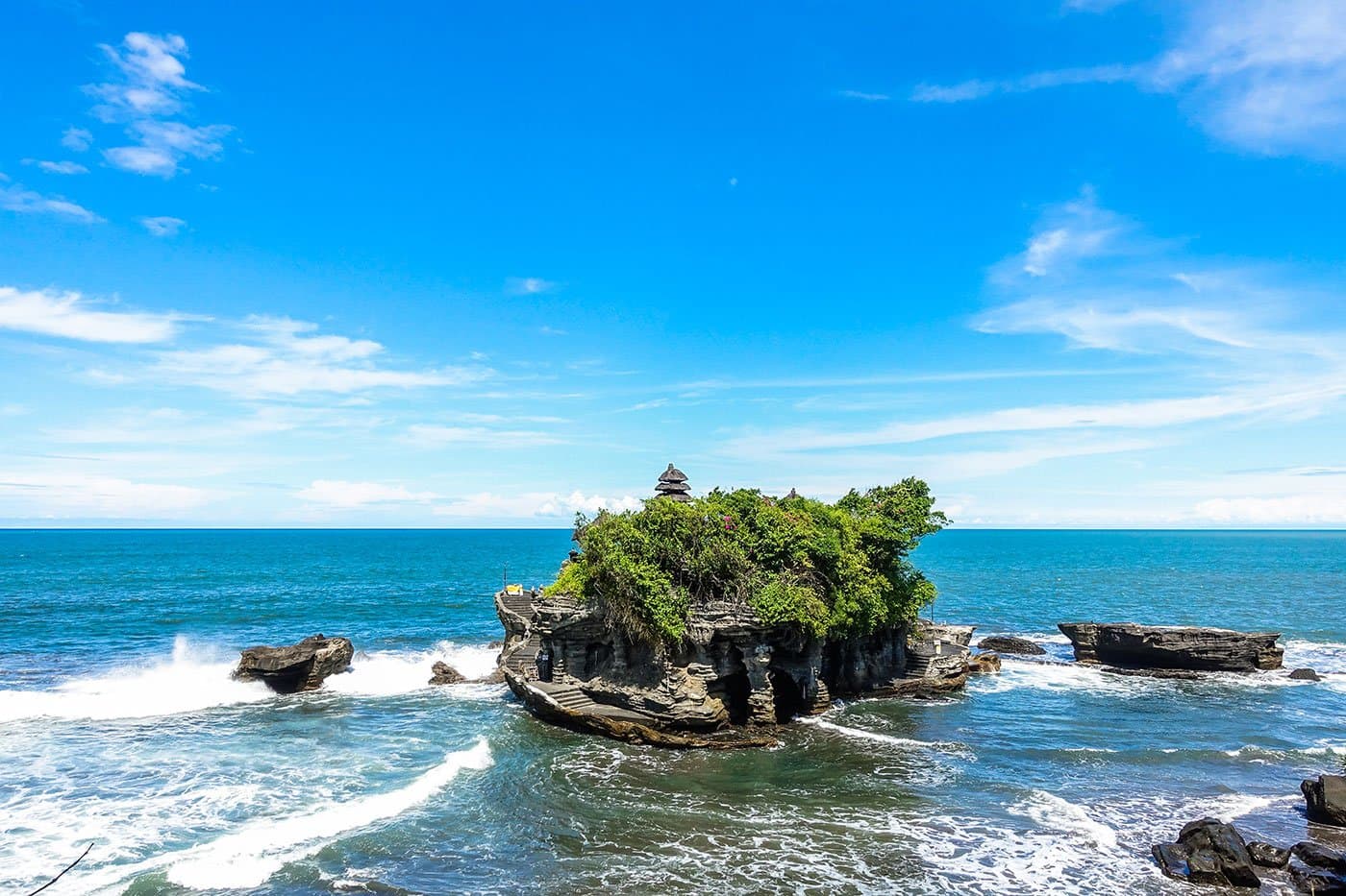Rocky sea outcrop topped with green shrubs and a small temple roof, waves breaking against it under a blue sky