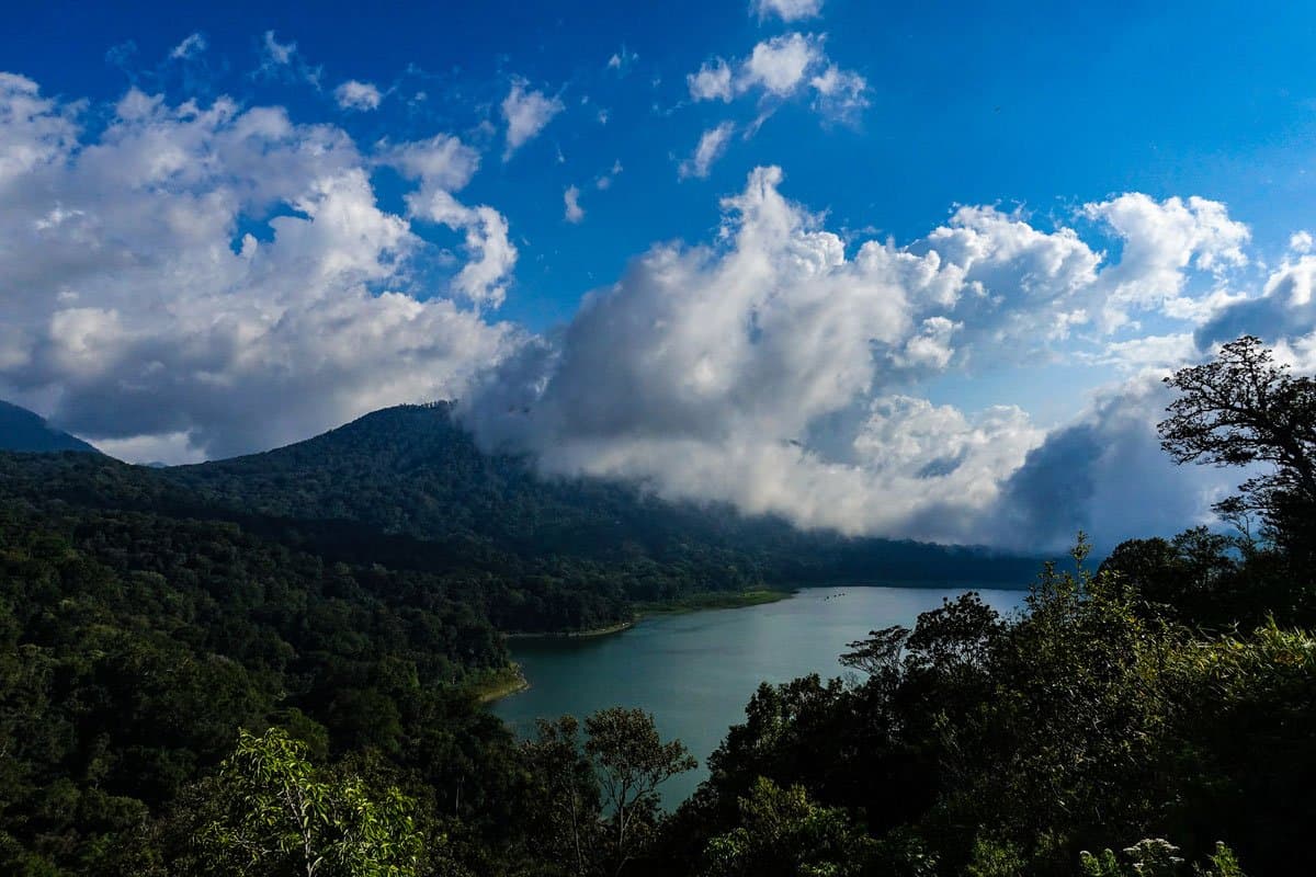 Elevated view of a calm lake surrounded by forested hills with clouds drifting through the valley below a blue sky