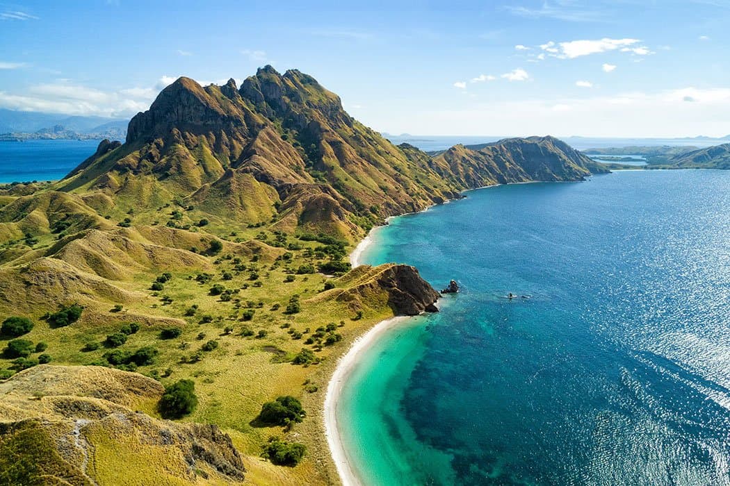 Aerial view of rugged green hills meeting a turquoise bay with two swimmers visible near the shoreline