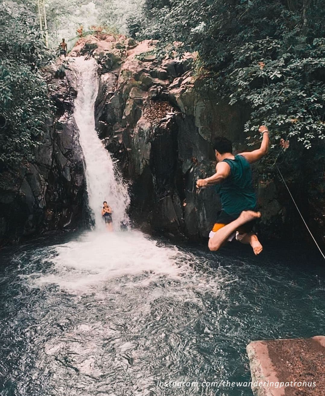 A person leaping into a river pool below a waterfall, with another swimmer standing beneath the falling water