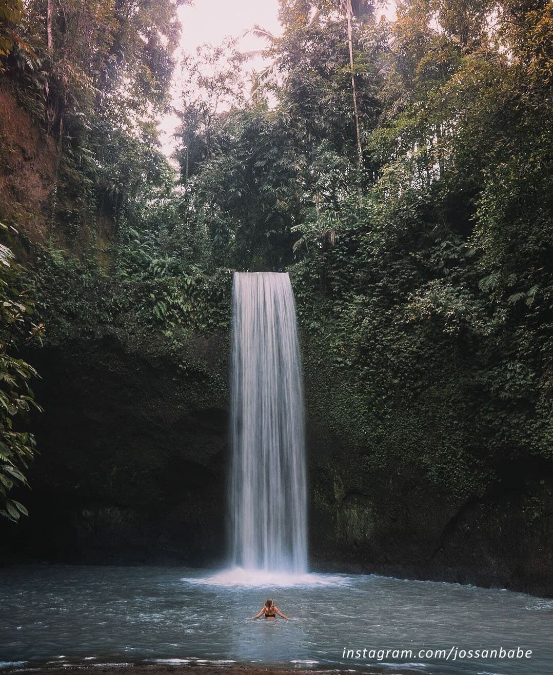 A tall narrow waterfall drops into a pool surrounded by dense jungle, with a swimmer standing in the water