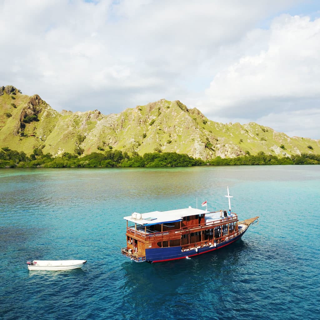 Sokaraja liveaboard anchored in turquoise water near a small dinghy, with green hills in the background