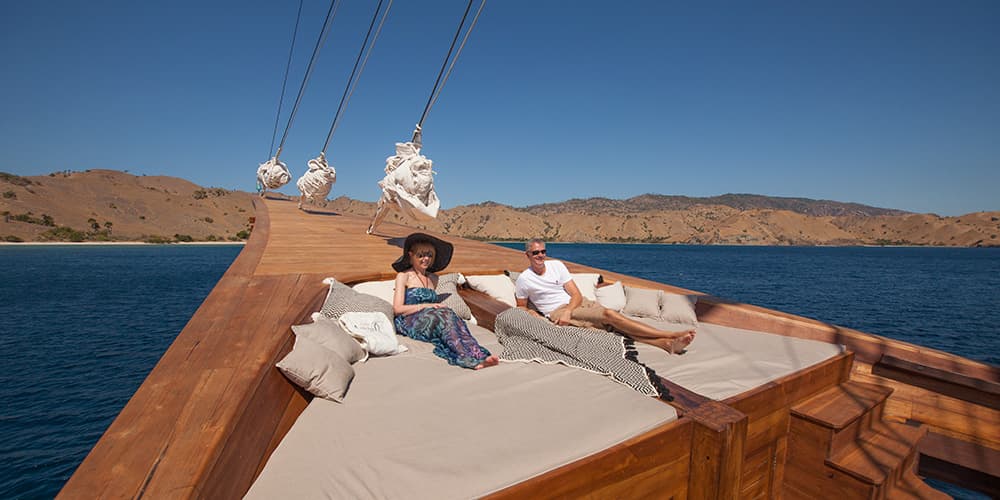 Two guests relax on the bow cushions of the Prana with hills and calm blue water in the background