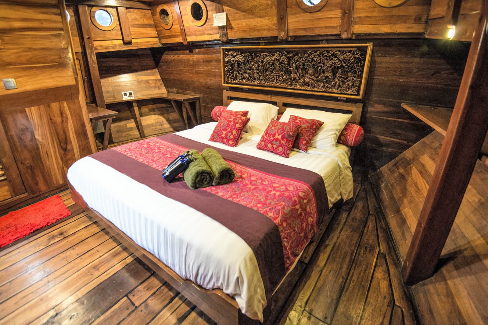 Wood-paneled cabin aboard Ilike Liveaboard with a double bed, red patterned cushions, rolled towels and portholes above