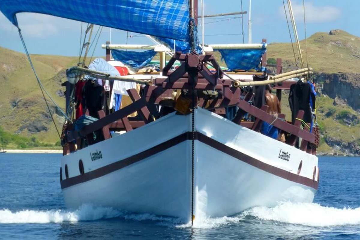Bow view of the wooden liveaboard Lambo with blue sails, wetsuits drying on deck, hills in the background