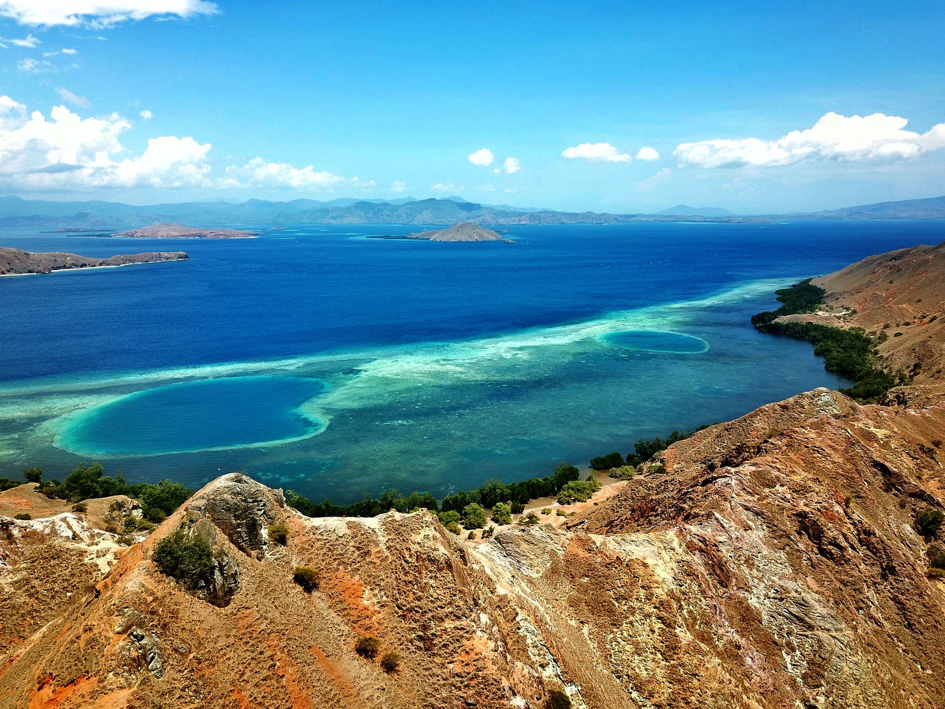 Aerial view of arid hills overlooking a curved reef and turquoise shallows meeting deep blue sea, distant mountains under a partly cloudy sky