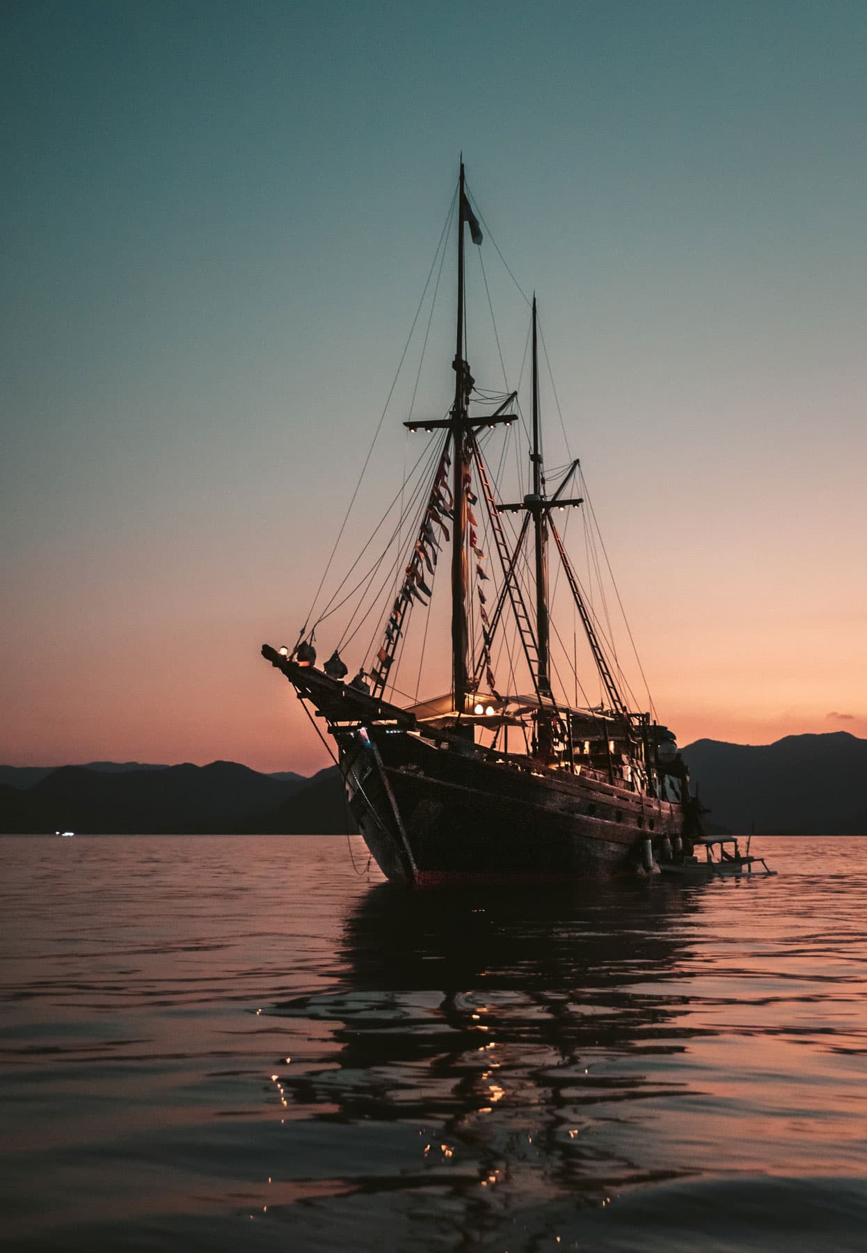 Two-masted wooden liveaboard anchored on calm water at sunset with hills silhouetted along the horizon