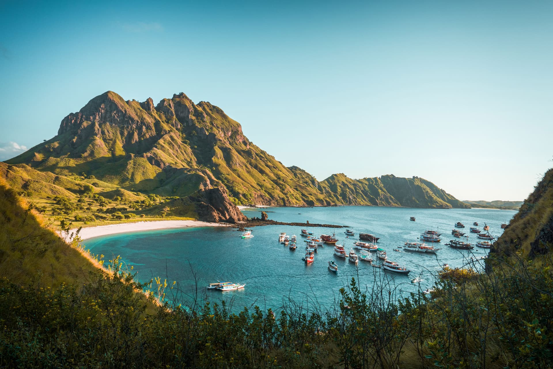 Boats anchored in a calm bay beside a white sand beach, framed by rugged green hills at sunset