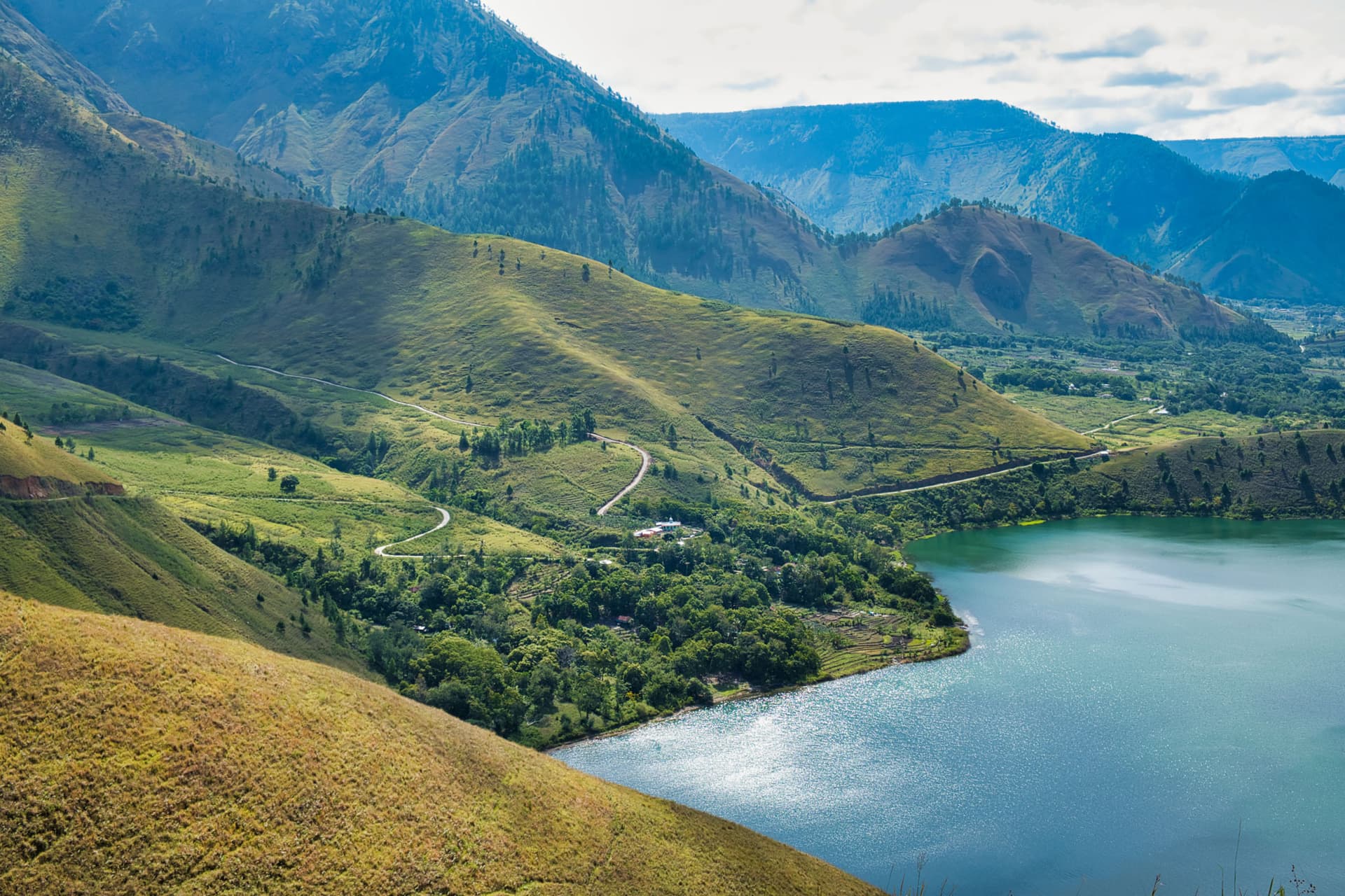 Aerial view of a winding road through green hills leading down to a lake surrounded by mountains