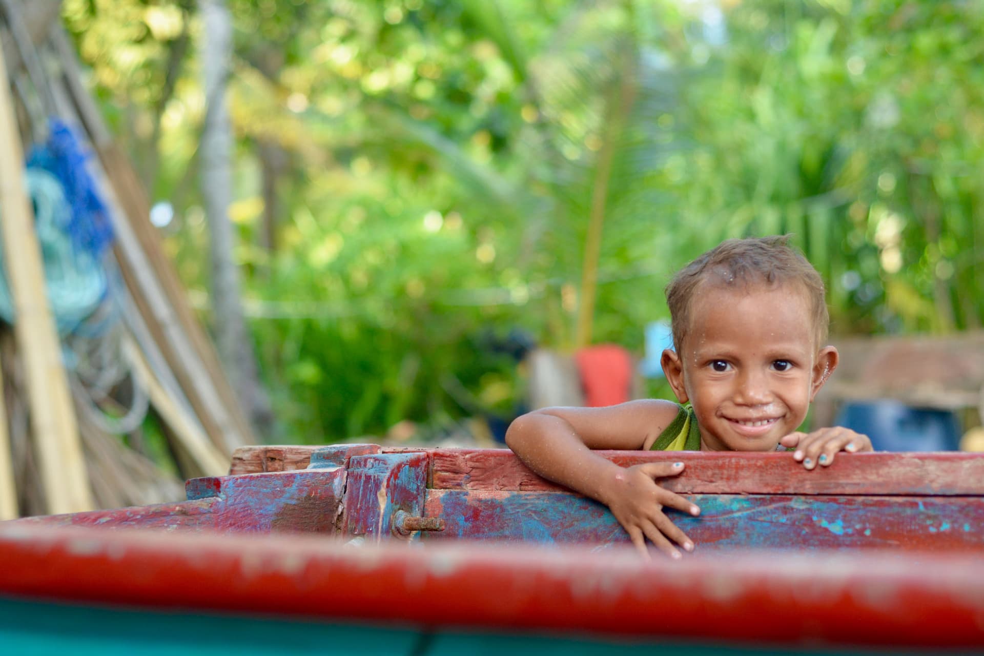 A child smiles while peeking over the edge of a weathered red wooden boat, surrounded by green foliage