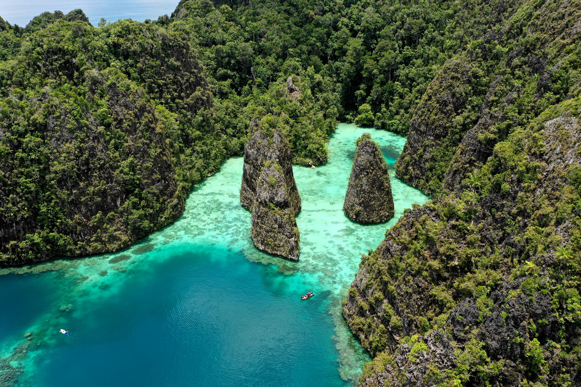 Aerial view of limestone karst peaks rising from a turquoise lagoon surrounded by dense forested cliffs, with a small boat below