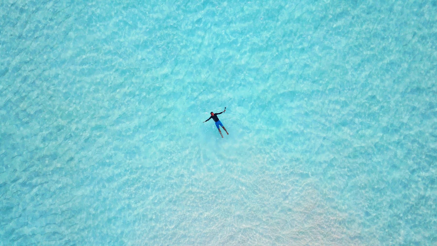 Aerial view of a swimmer floating on turquoise water, arms outstretched, holding a selfie stick