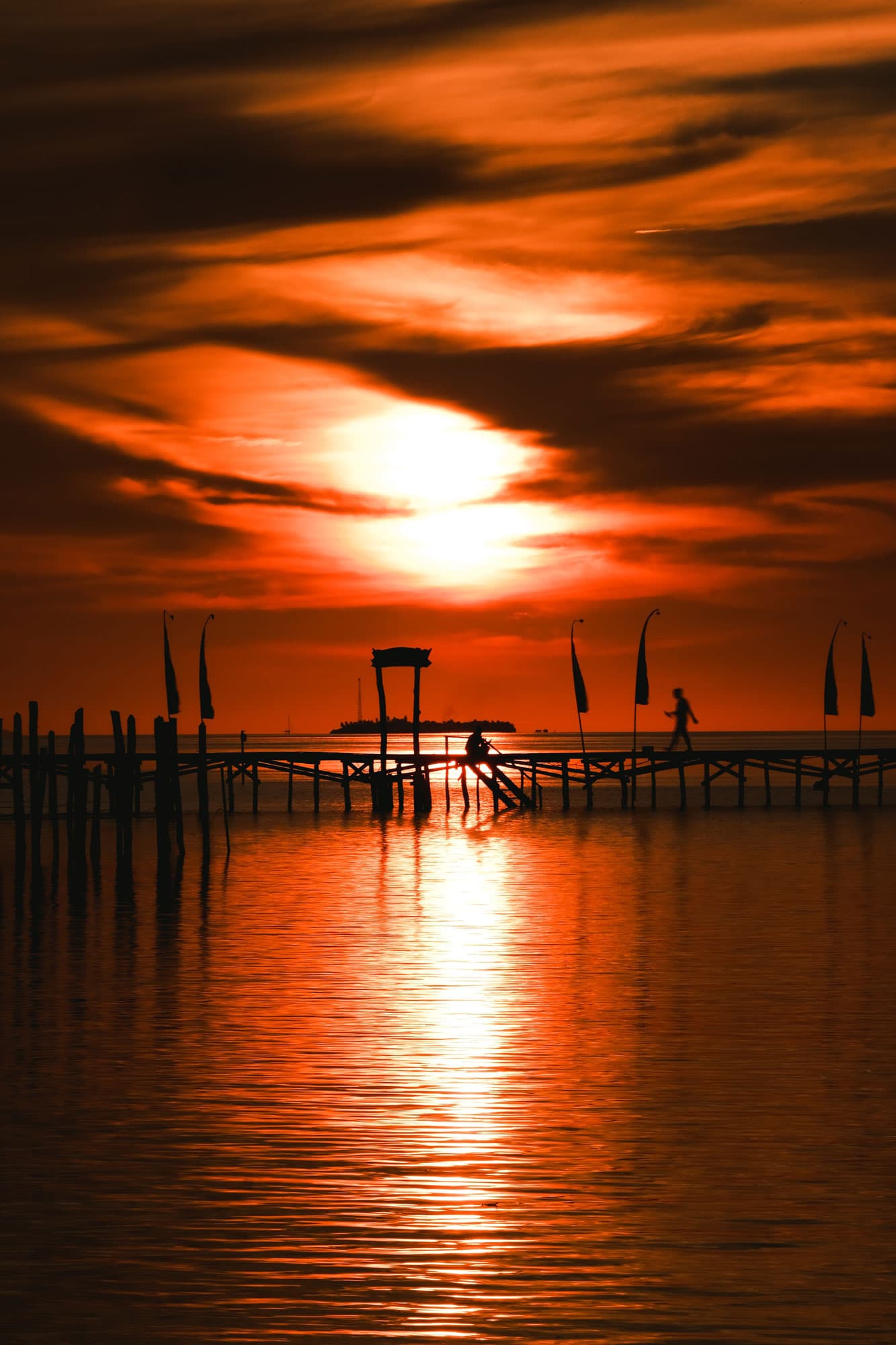 Silhouetted pier with flags and a person walking, set against a deep orange sunset reflected on calm water