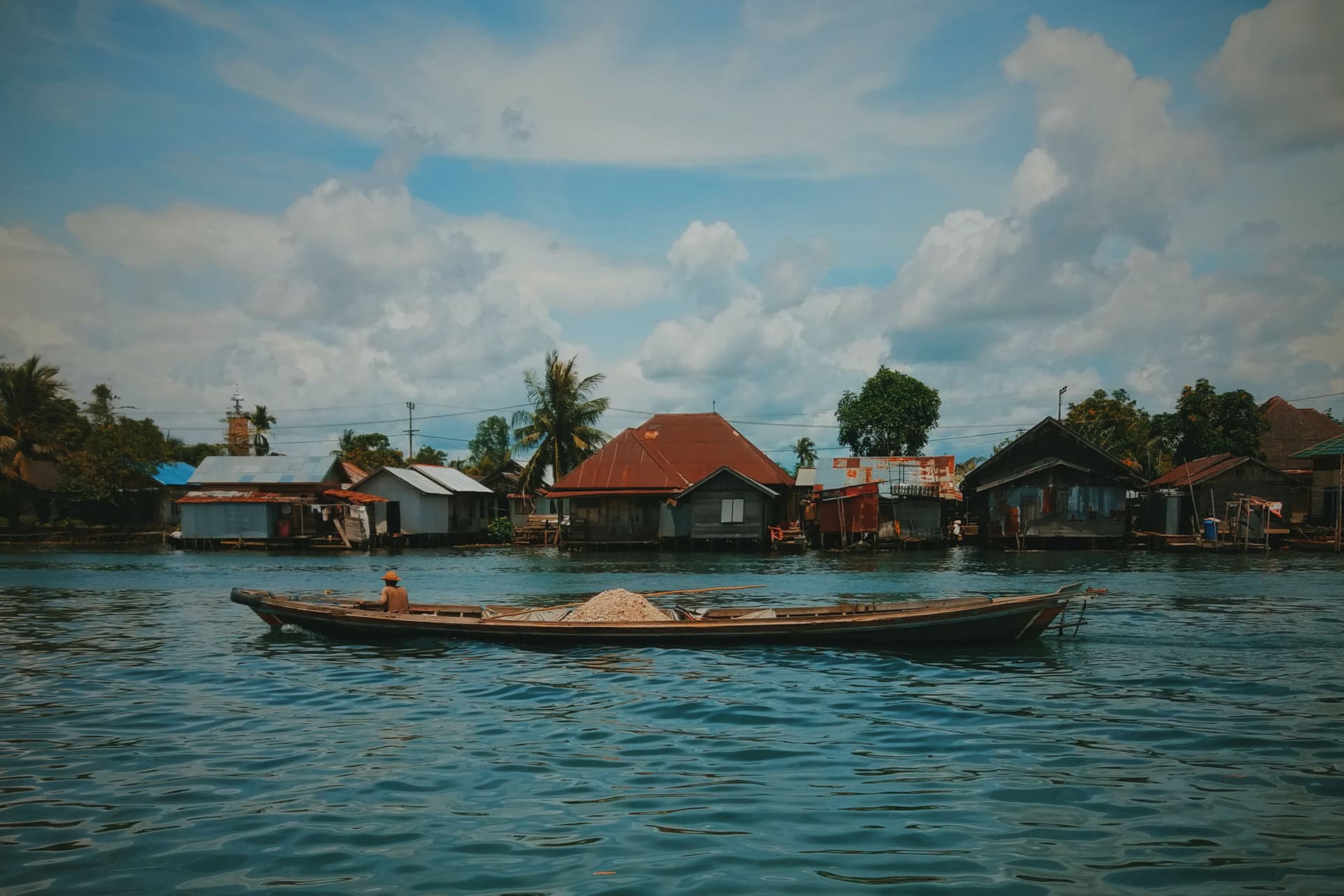 A wooden boat carrying gravel glides past a row of stilt houses along a river under a partly cloudy sky