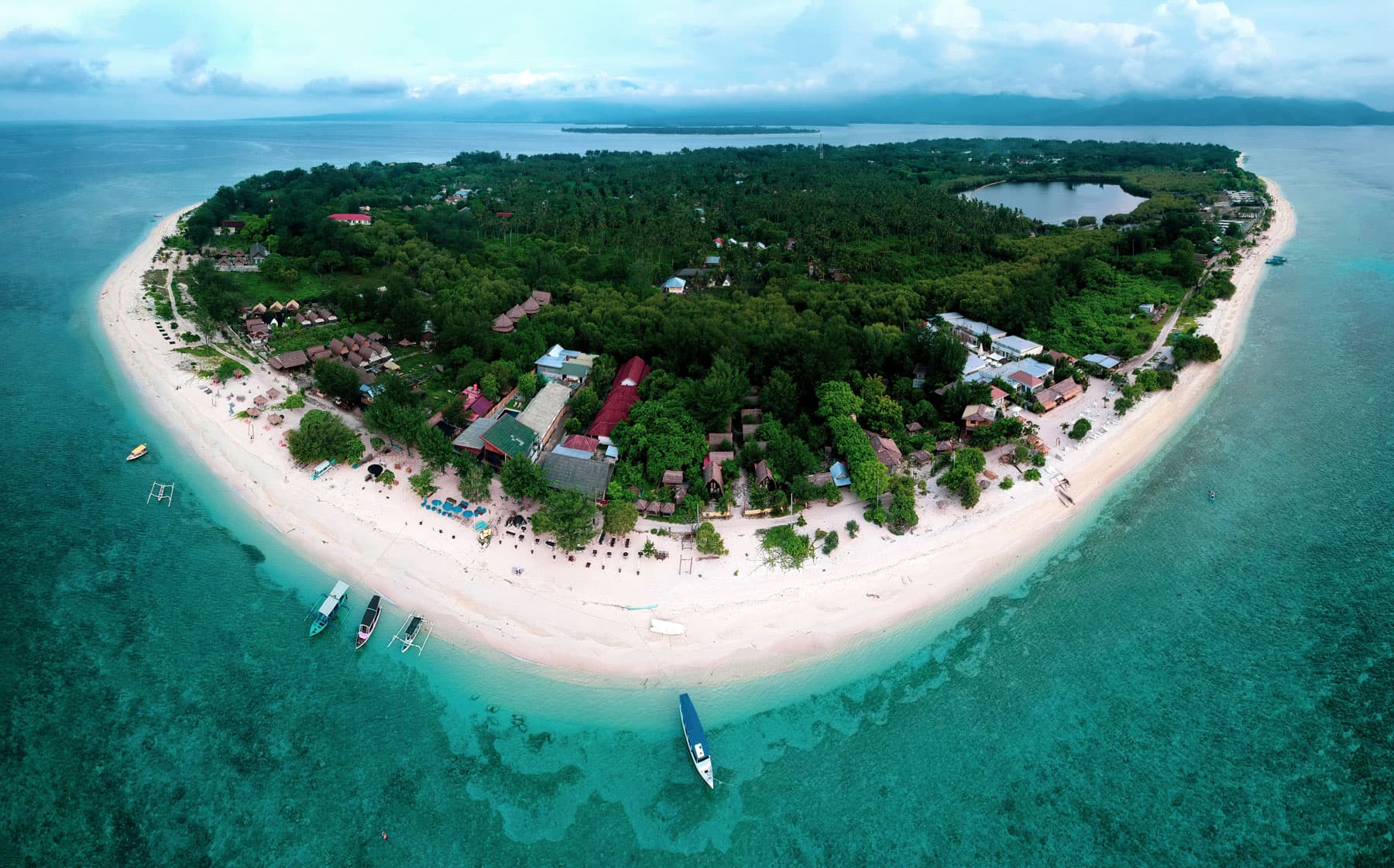 Aerial view of a small tropical island with dense forest, beachfront buildings, a sandy shoreline and boats anchored in turquoise water