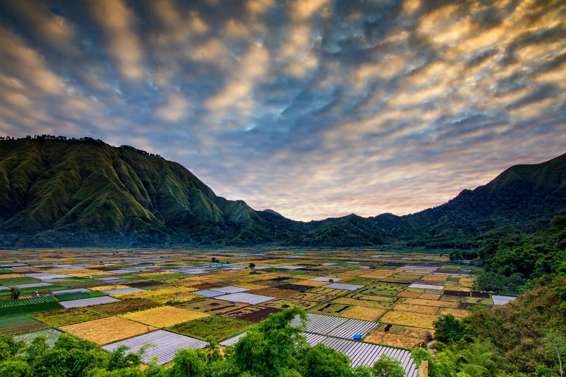 Patchwork of farm fields in a valley surrounded by green mountains under a dramatic clouded sky at sunset