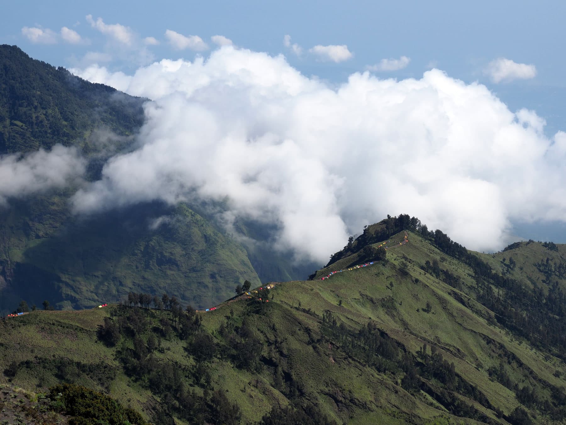 Green mountain ridge with a line of small tents and colorful flags along its crest, surrounded by thick clouds