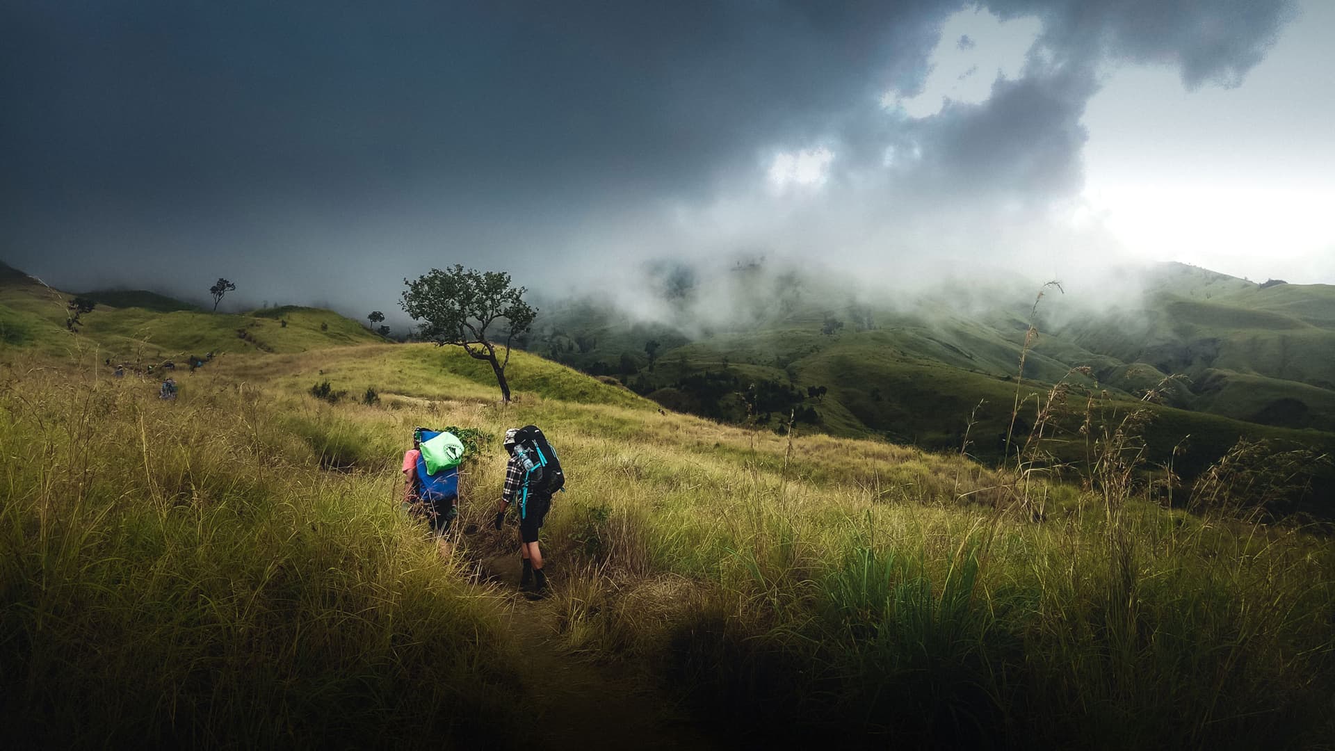 Two hikers with backpacks walking a grassy hillside trail beneath dark storm clouds and low mist
