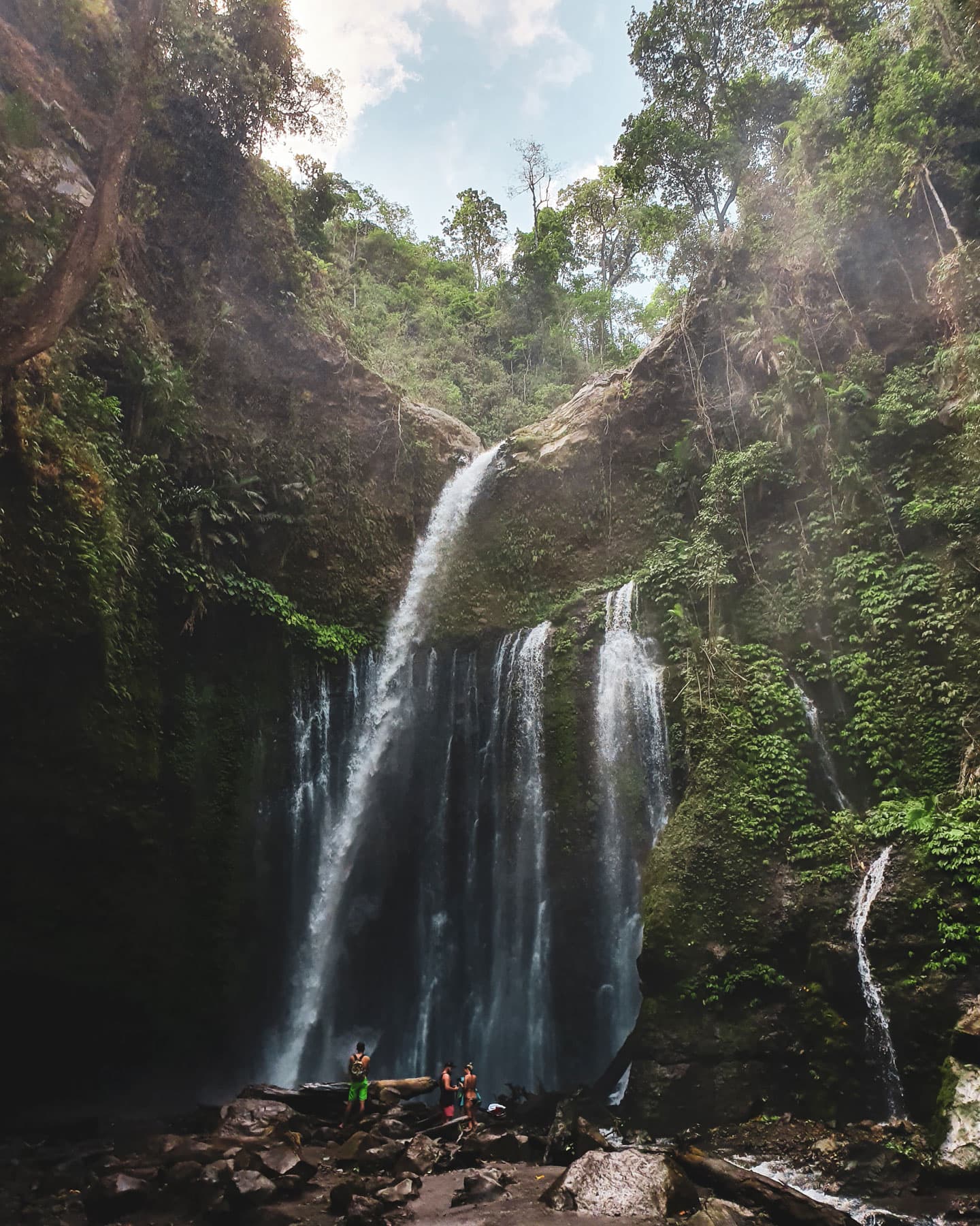 Tall waterfall dropping over a jungle cliff into a rocky pool, with small figures standing at its base