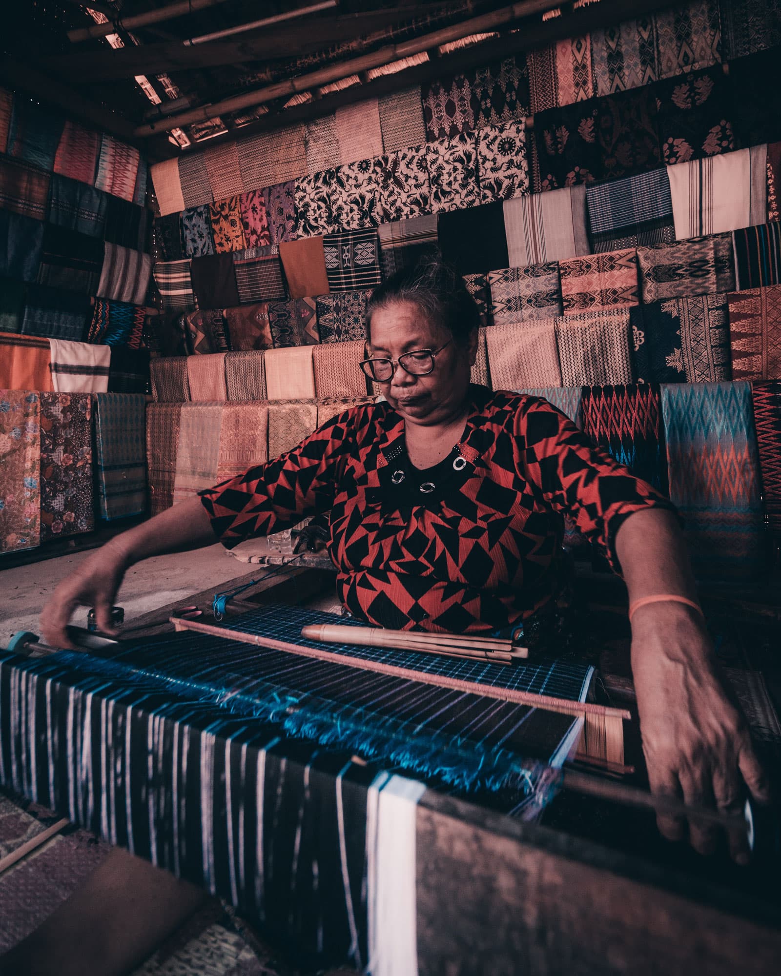 Woman weaving on a traditional loom, surrounded by stacks of colorful handwoven textiles on the walls