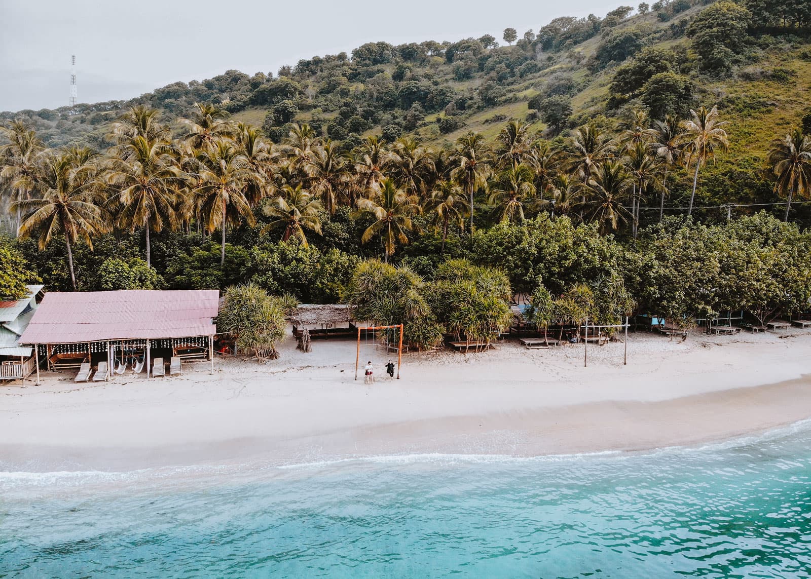 Aerial view of a beach hut and swings on white sand beside turquoise water, backed by palm trees and a forested hillside