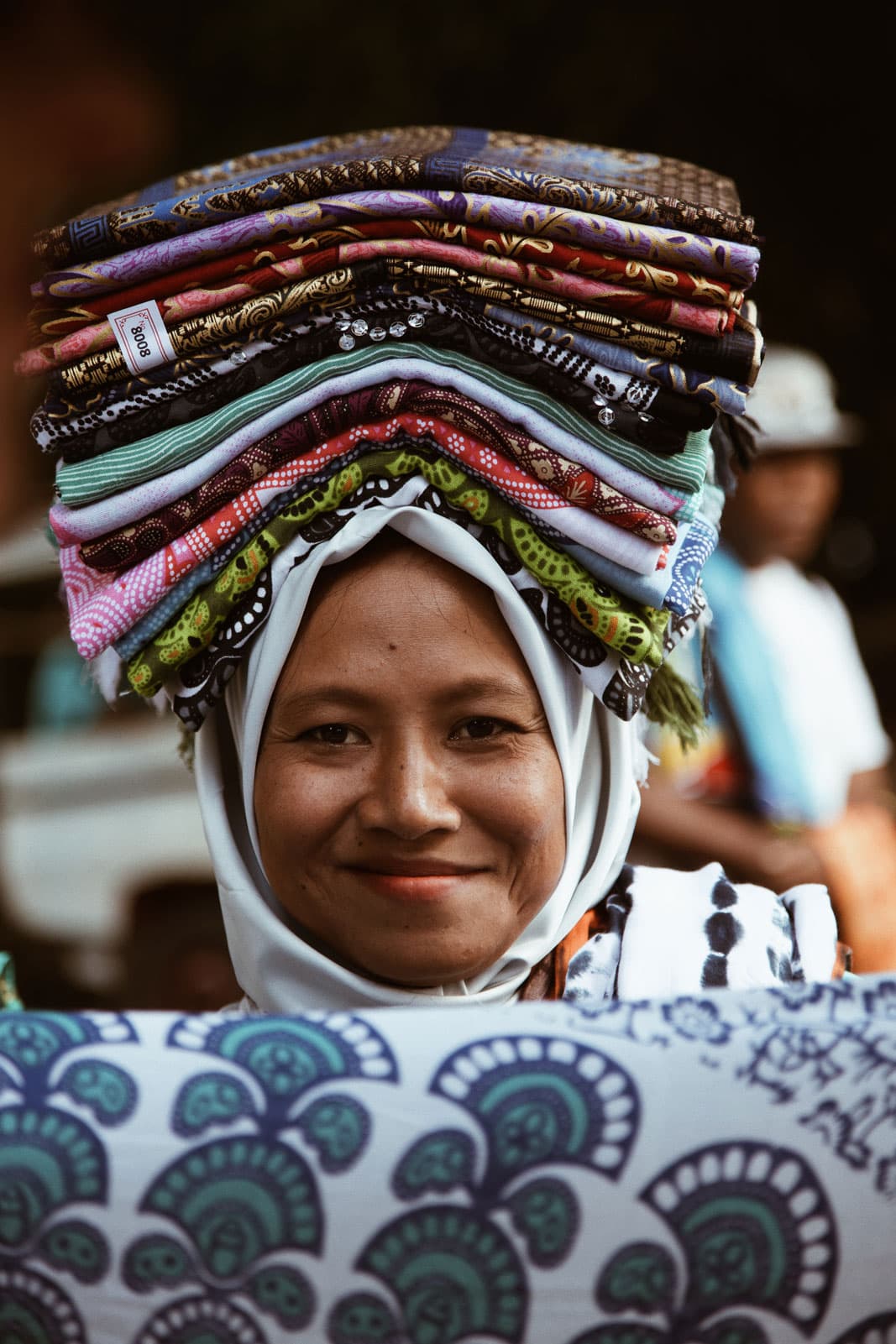 Smiling woman balancing a tall stack of folded batik textiles and sarongs on her head