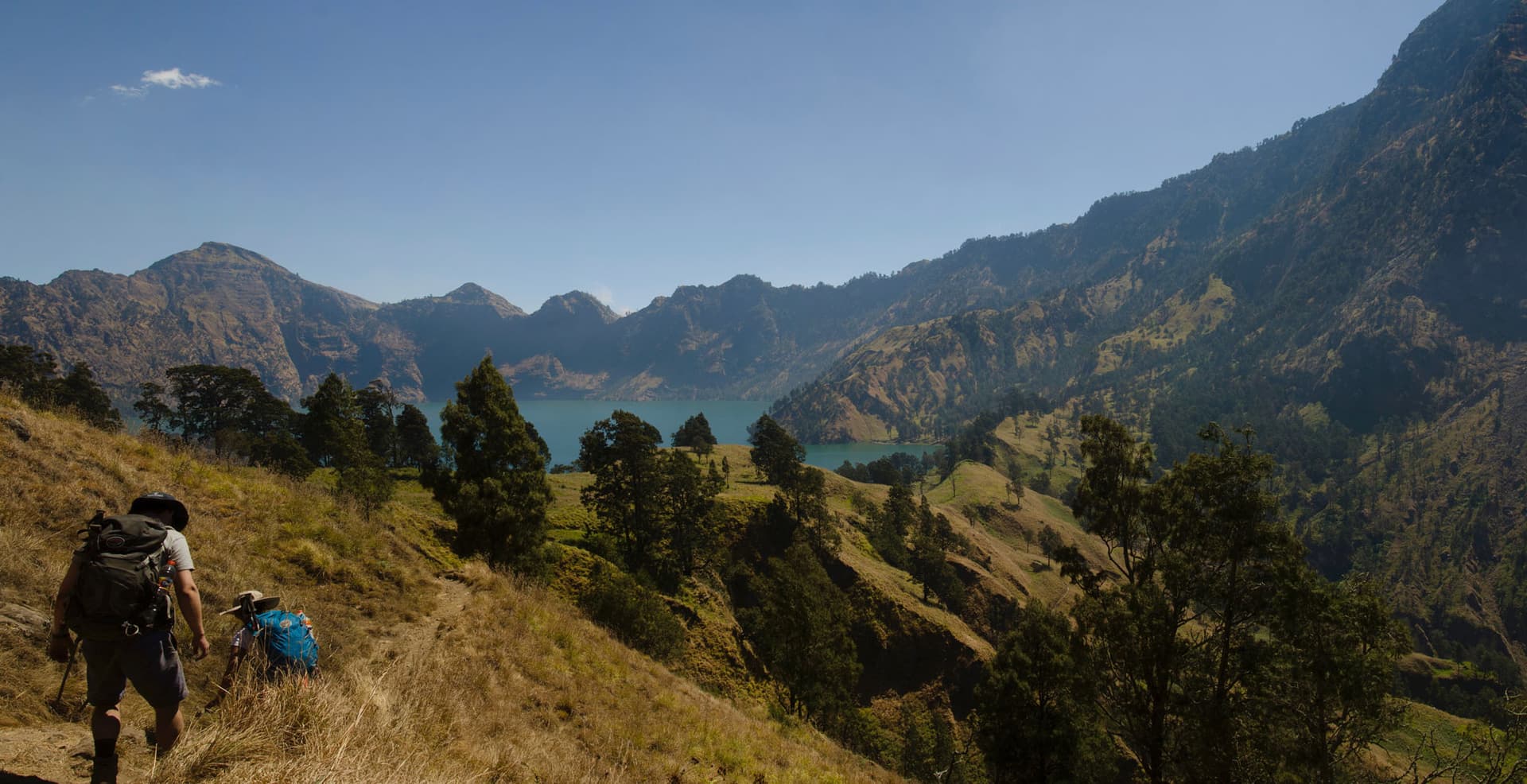 Hikers with backpacks descending a grassy ridge trail overlooking a turquoise crater lake surrounded by mountains