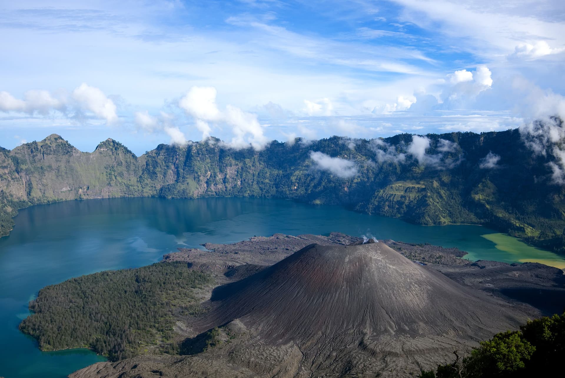 Aerial view of a small cinder cone with a smoking crater inside a larger volcanic caldera lake ringed by forested cliffs