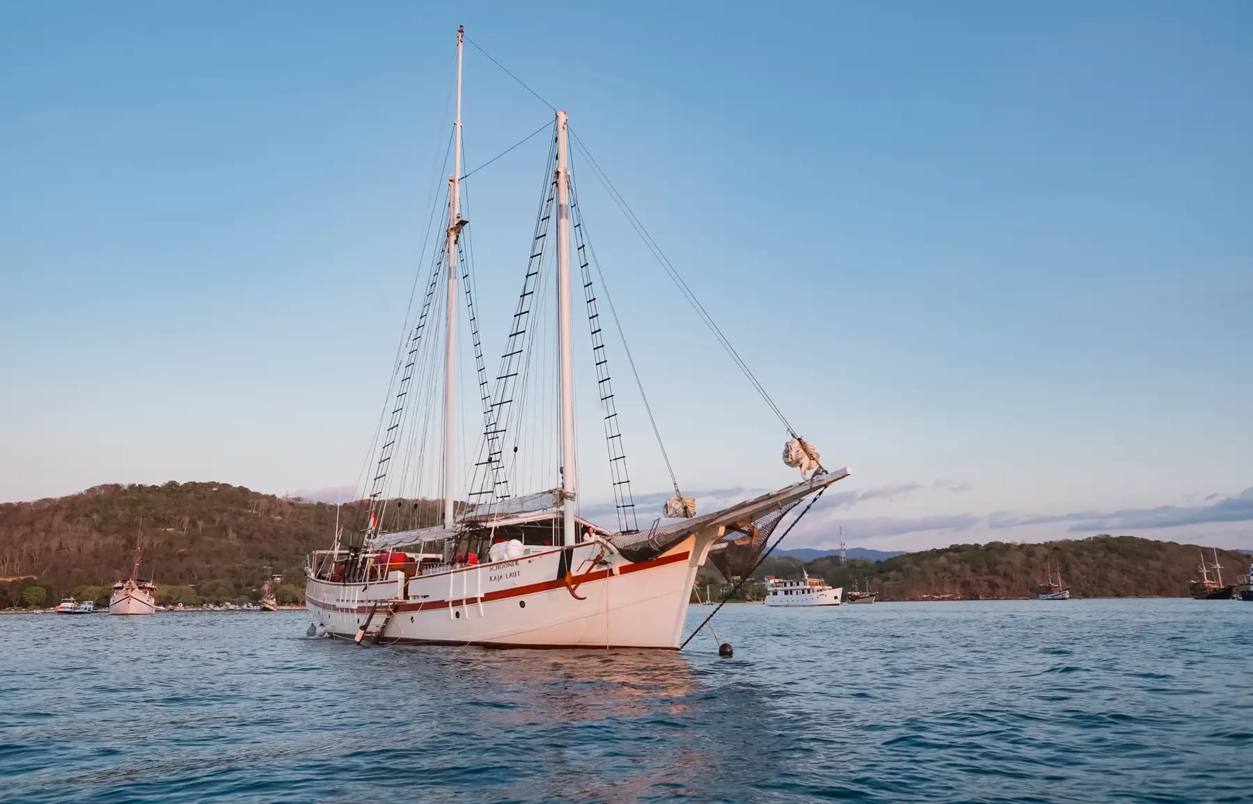 Emperor Raja Laut liveaboard with two tall masts anchored near a coastline at dusk, other boats visible in the distance