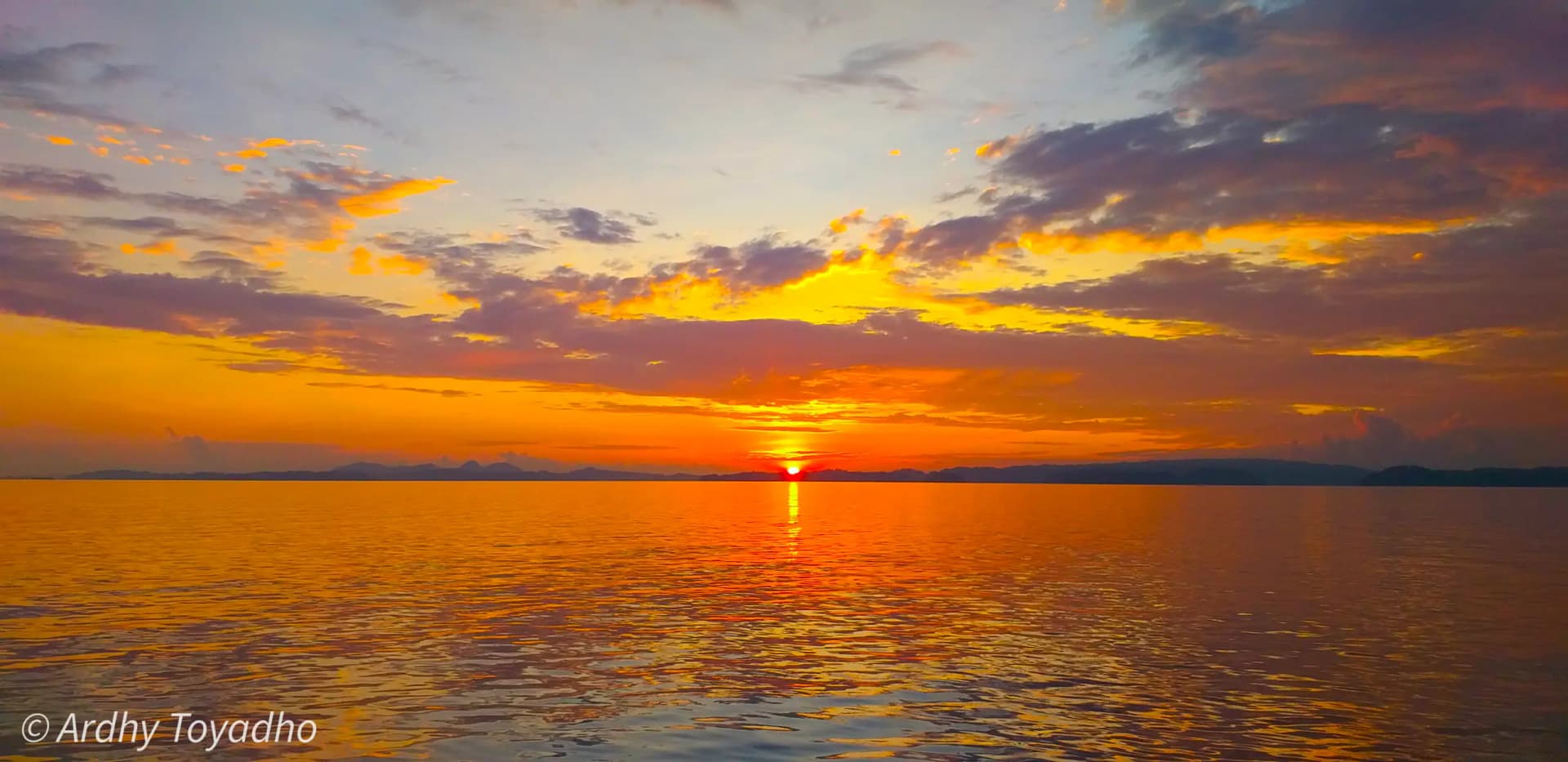 Vivid orange sunset over calm open water with layered clouds and a faint silhouette of distant hills on the horizon