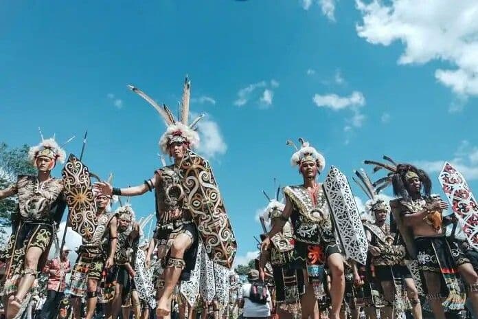 Men in traditional feathered headdresses and carved shields march in a procession under a blue sky with clouds
