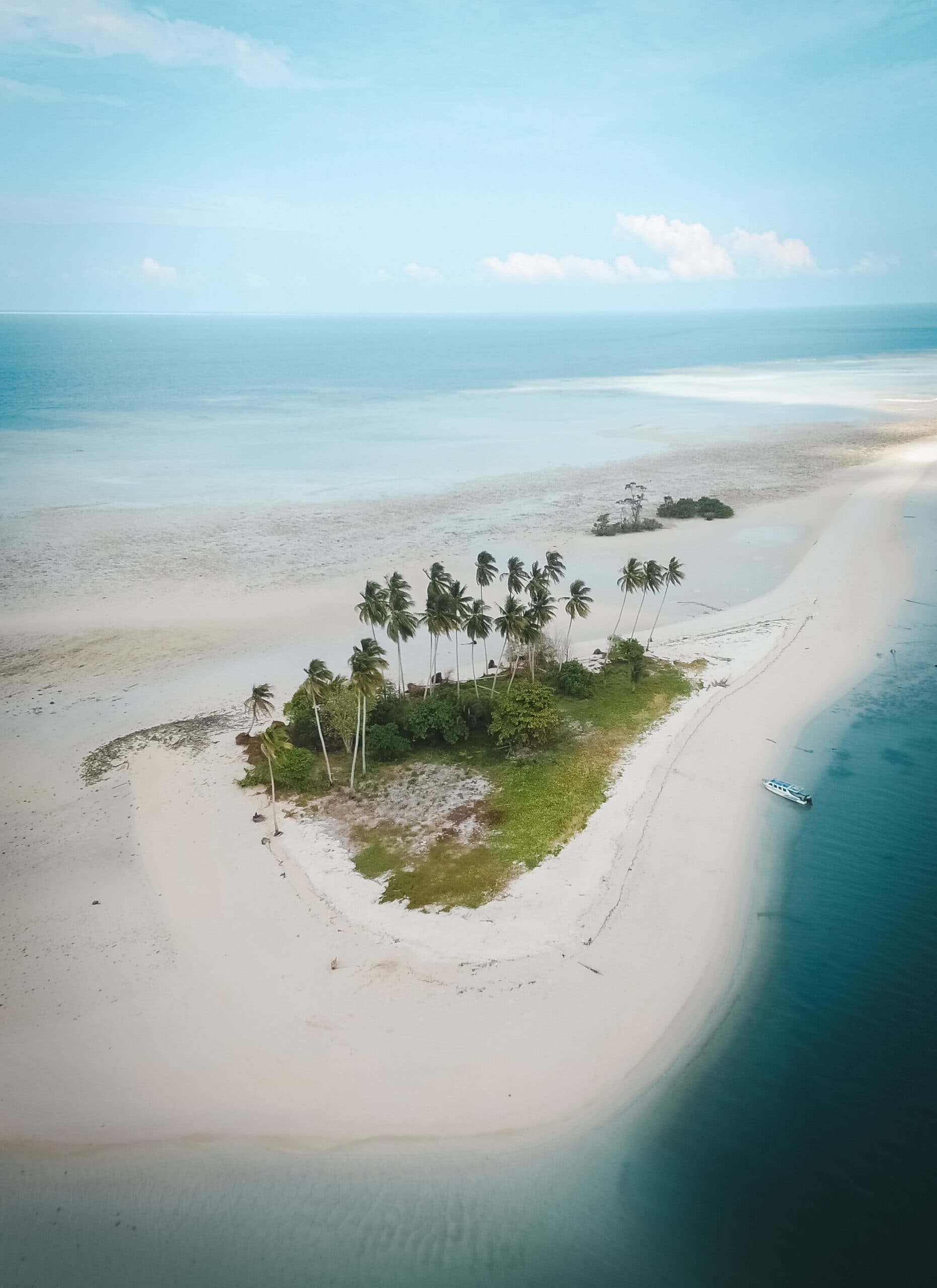 Aerial view of a small palm-covered sandy island surrounded by shallow turquoise water and a moored boat