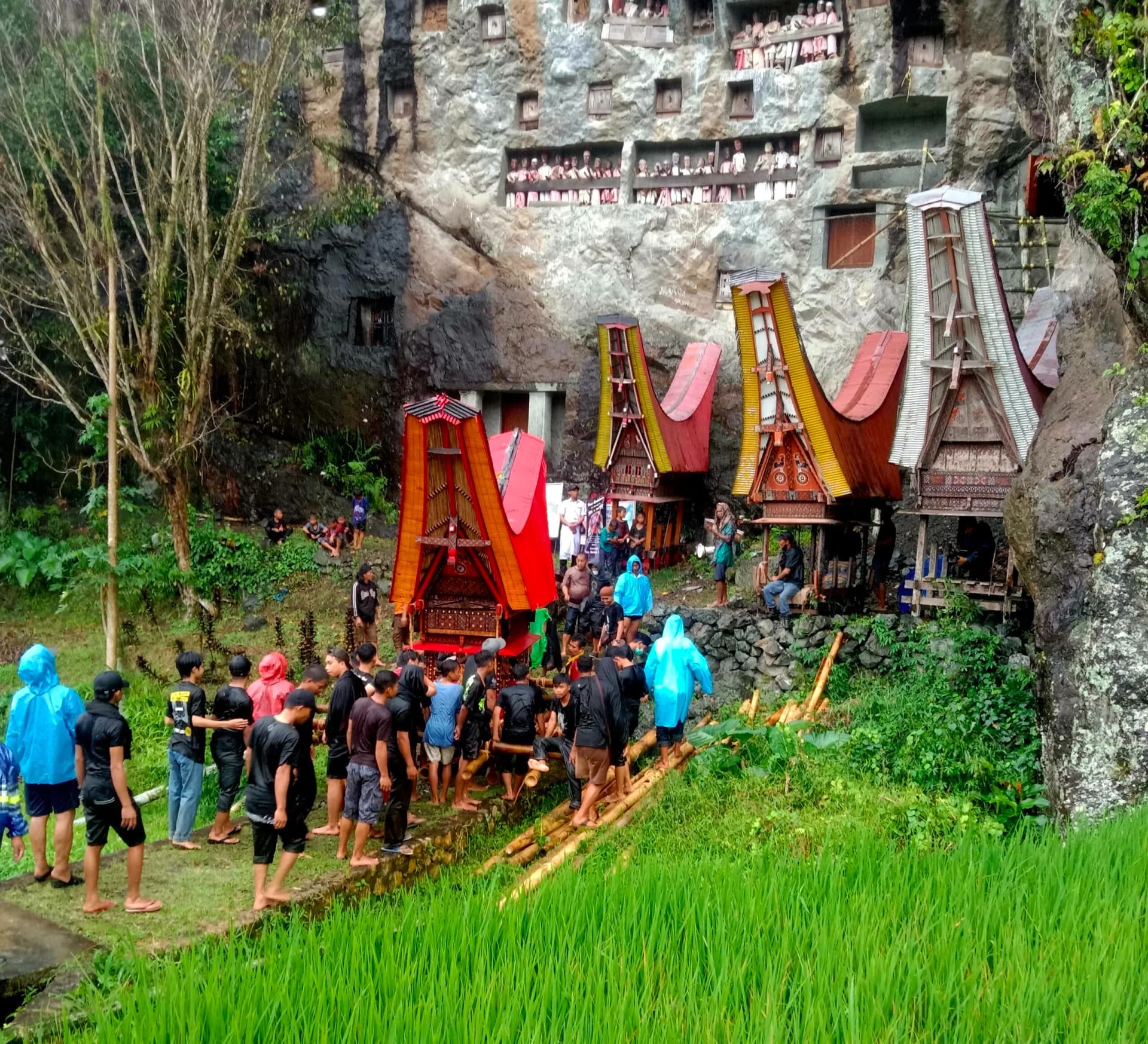 Group carrying a decorated funeral tower toward a cliffside burial site with carved wooden structures and effigies