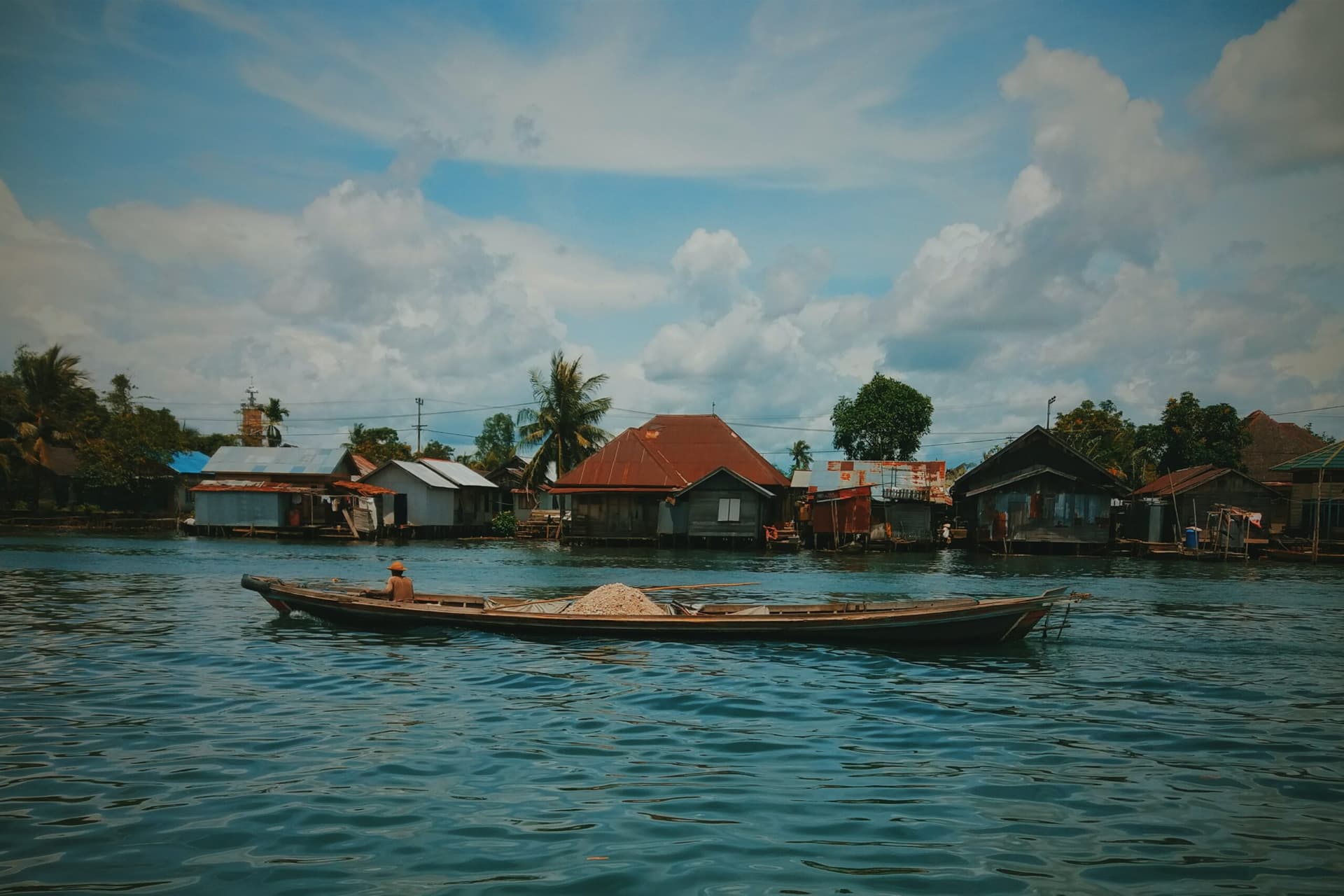 Wooden boat loaded with gravel on a river, with stilt houses and palm trees lining the far bank