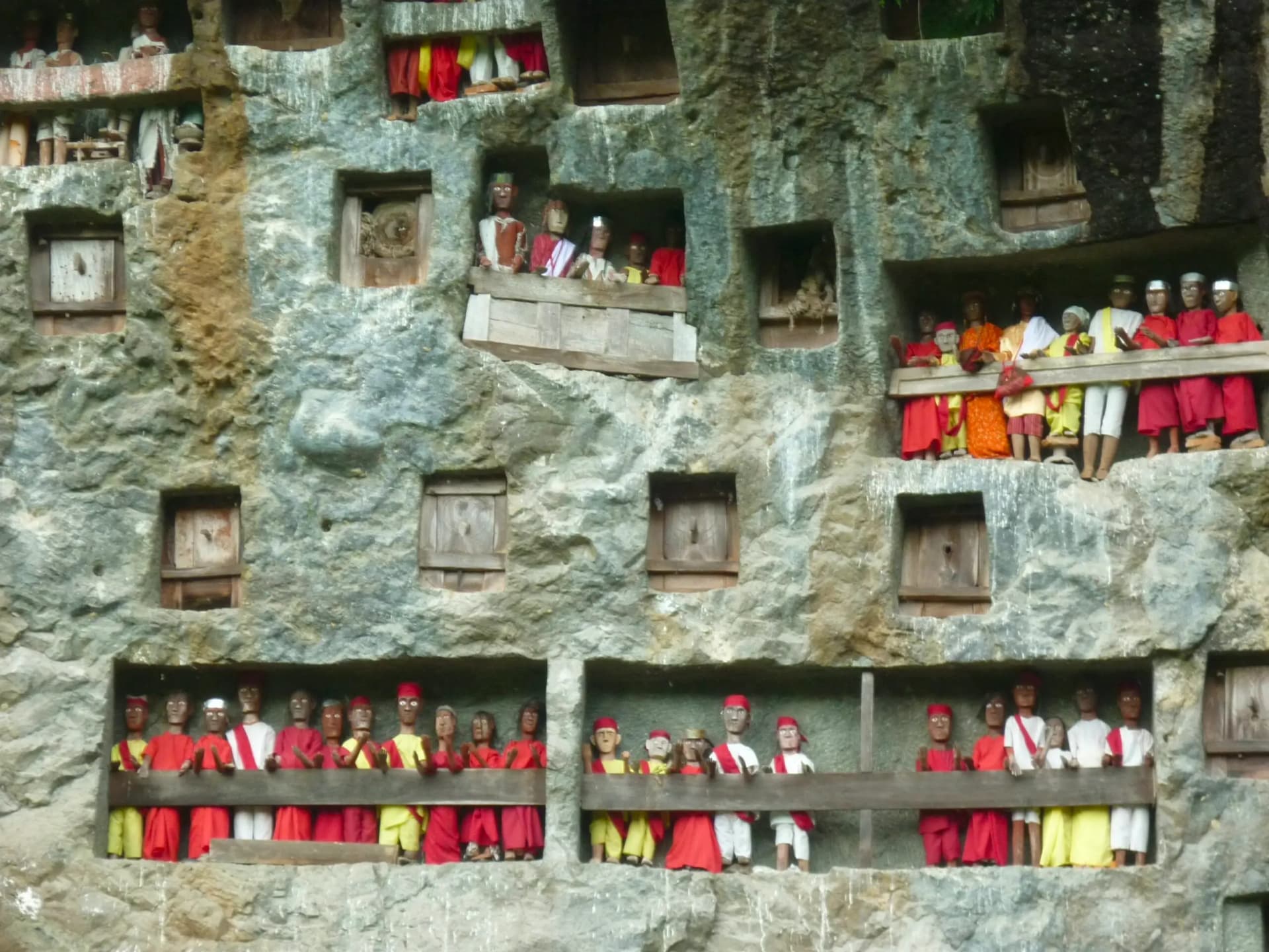 Rows of carved wooden effigies dressed in red, yellow and white displayed on balconies carved into a rocky cliff face