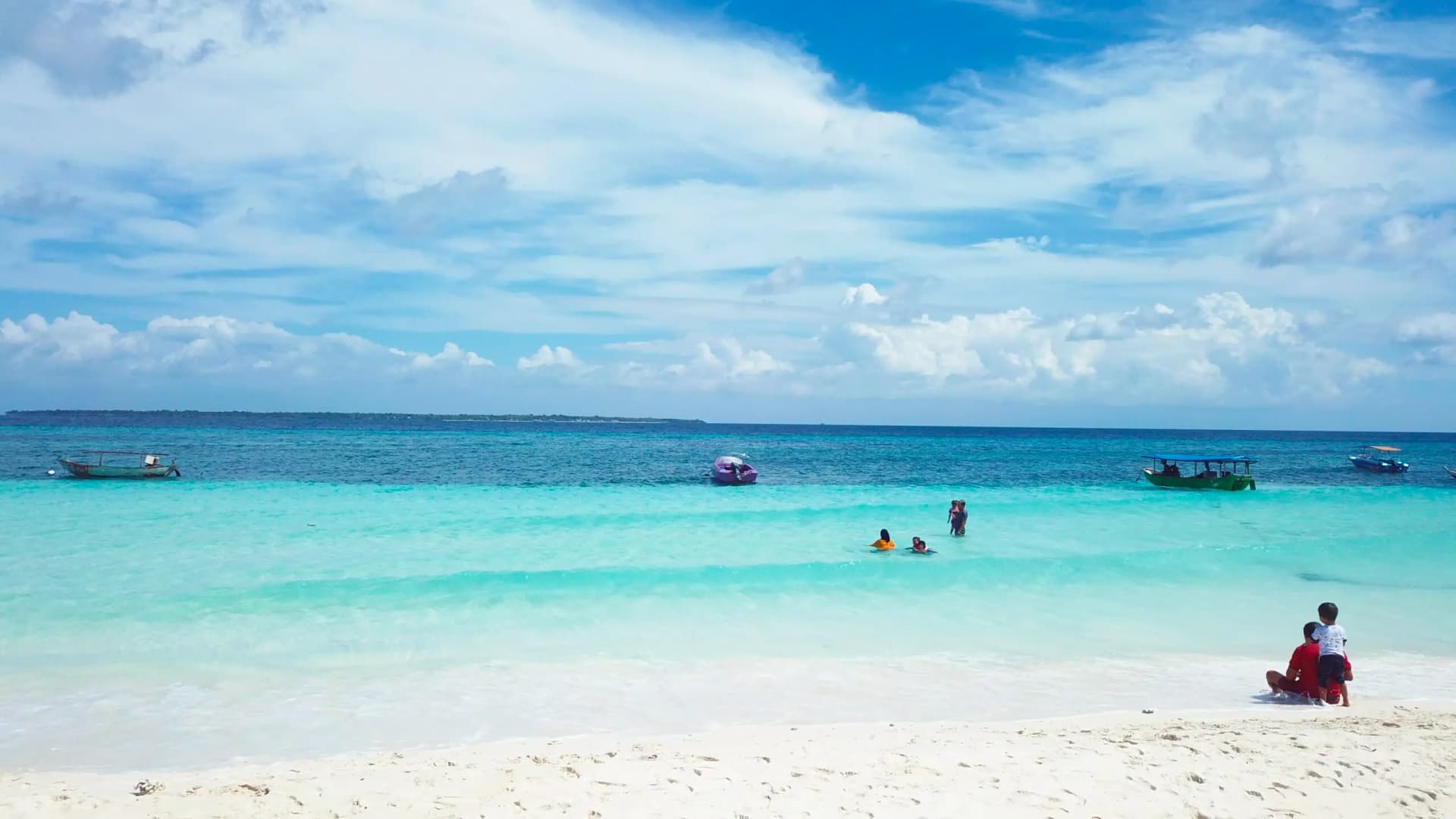Turquoise sea with small boats and swimmers, a person and child sitting on white sand under a partly cloudy sky