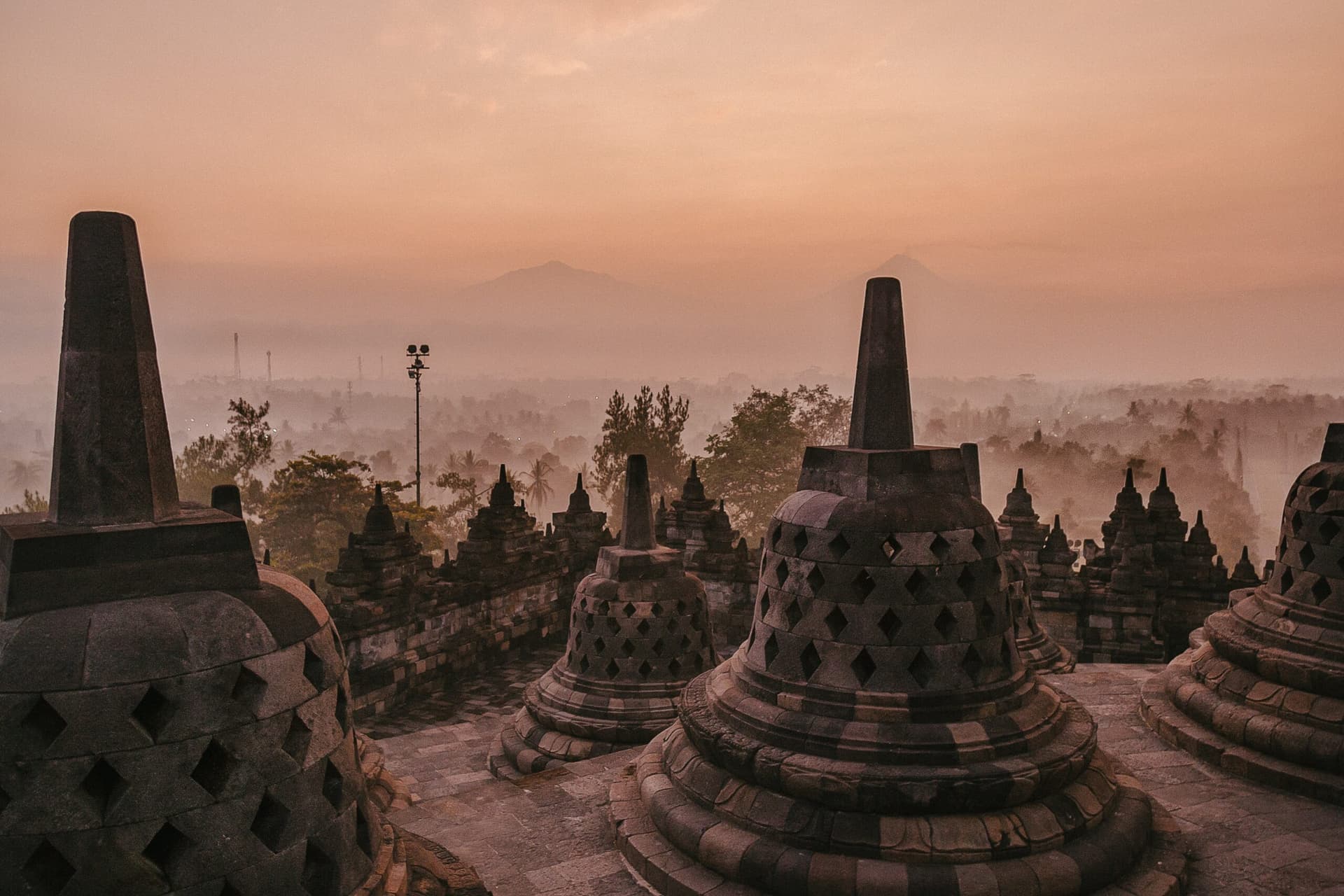 Stone stupas with latticed bell shapes on a temple rooftop overlooking misty treetops and distant hills at dawn