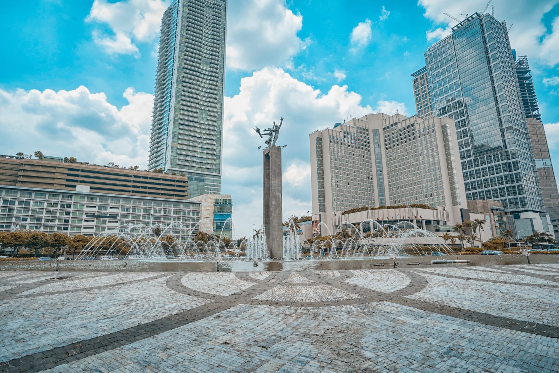 Selamat Datang Monument with fountains and surrounding skyscrapers on a paved plaza under a partly cloudy sky