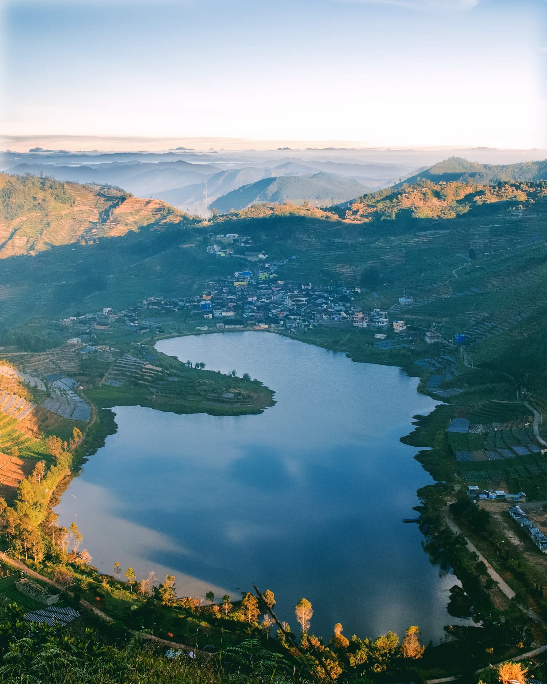 Aerial view of a heart-shaped lake surrounded by terraced hillsides, a small village, and layered mountains under a hazy sky