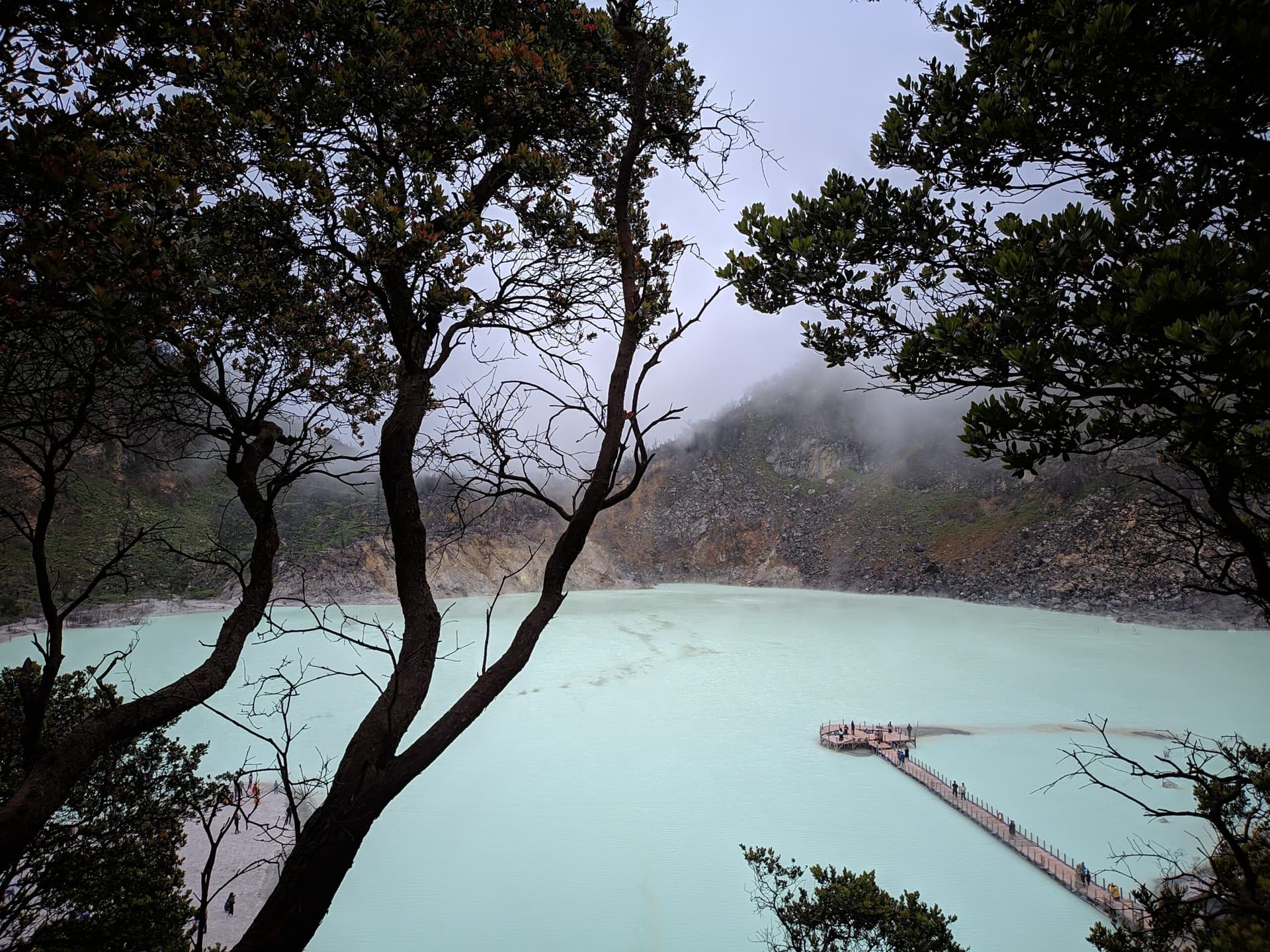 Milky turquoise crater lake seen through dark tree branches, with a wooden walkway and small figures near the shore
