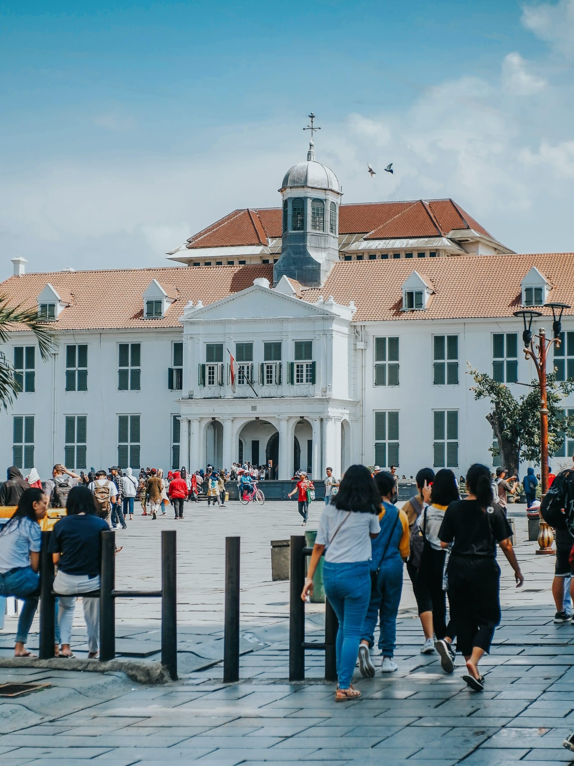Visitors walking across a paved square in front of a white colonial-era building with a clock tower and red tile roof