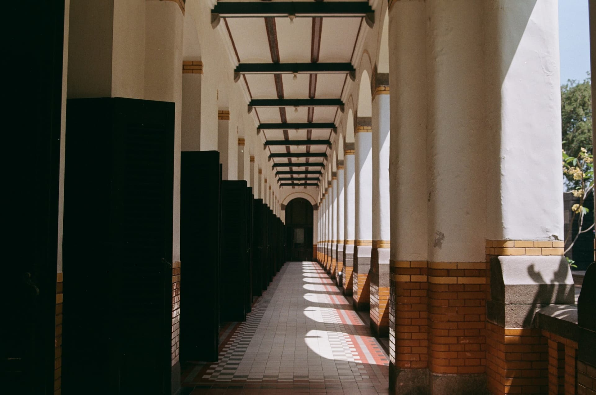 Long colonnaded corridor with arched columns, tiled floor, and dark wooden shutters casting striped shadows in sunlight
