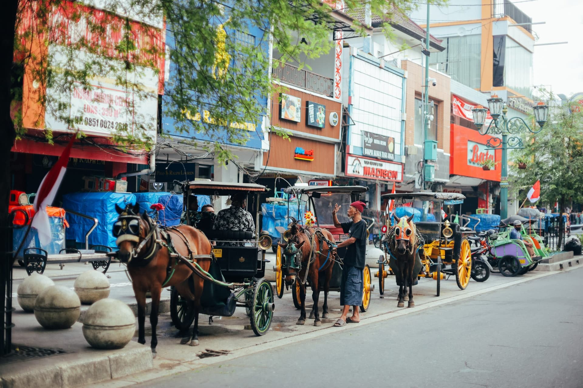 Horse-drawn carriages line a city street beside shopfronts, with a man standing between the horses