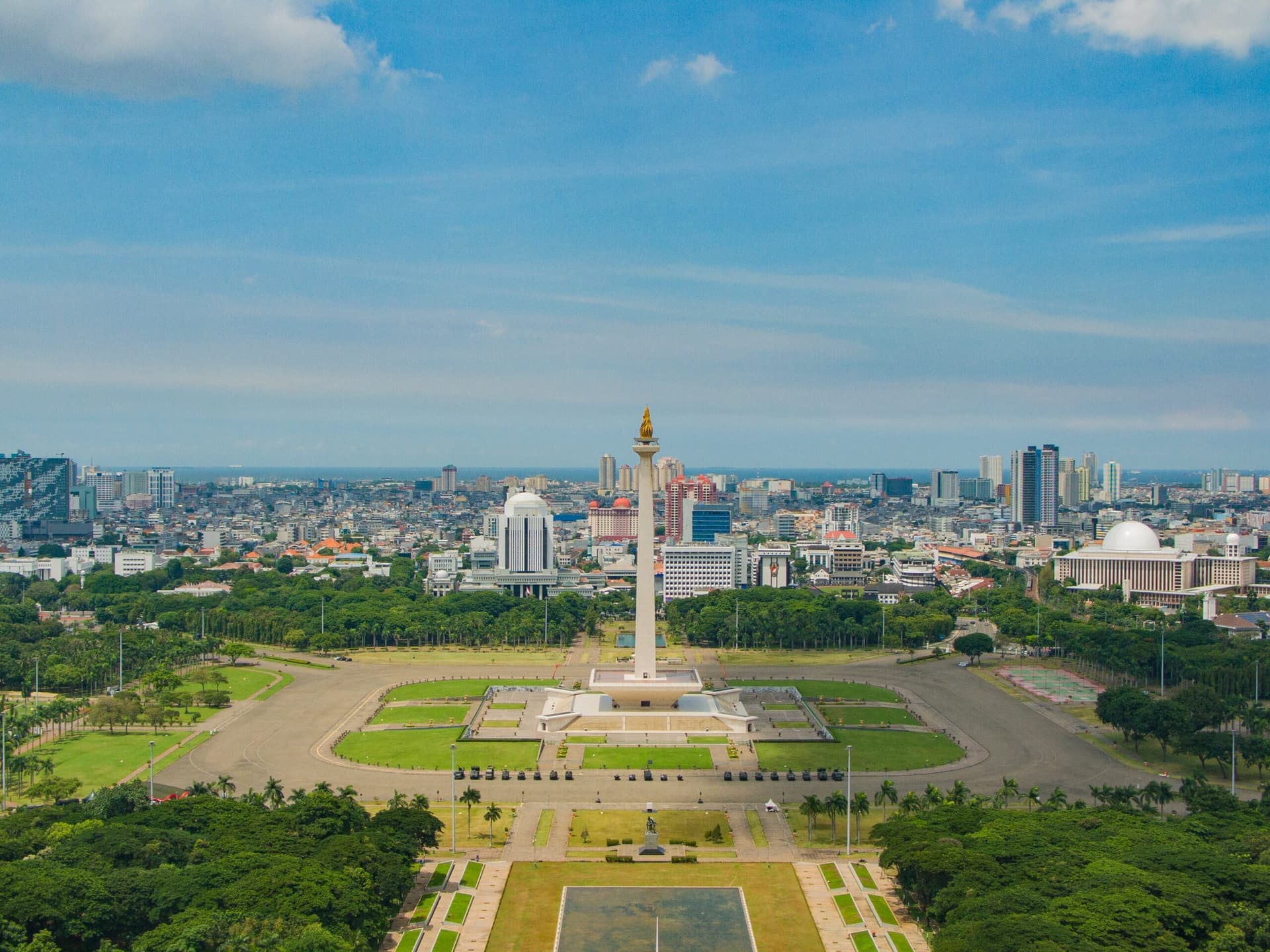 Aerial view of a city skyline with a tall monument topped by a flame, surrounded by parkland and tree-lined avenues