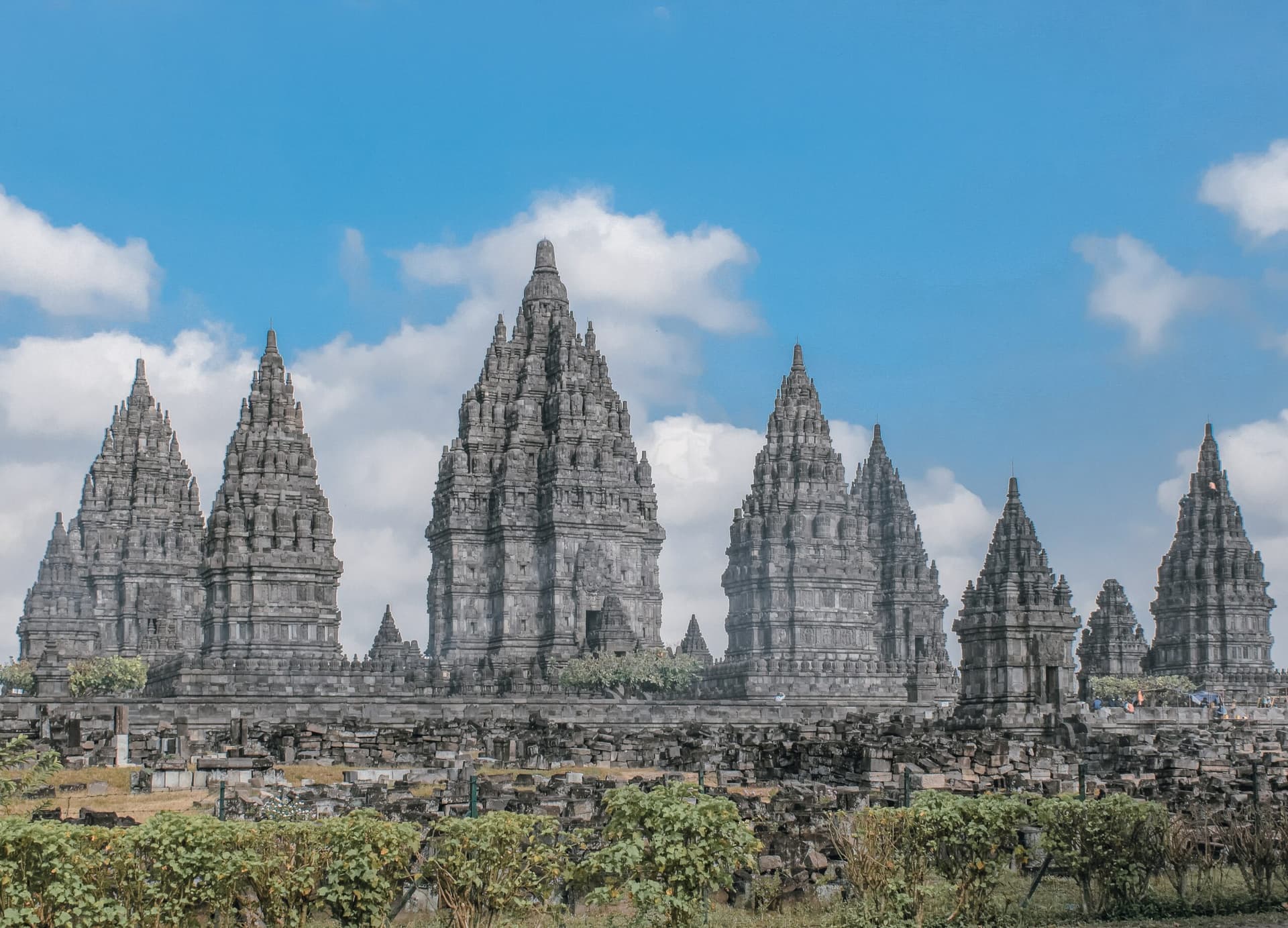Tall stone temple spires with intricate carvings rise against a blue sky with scattered clouds, ruins in the foreground