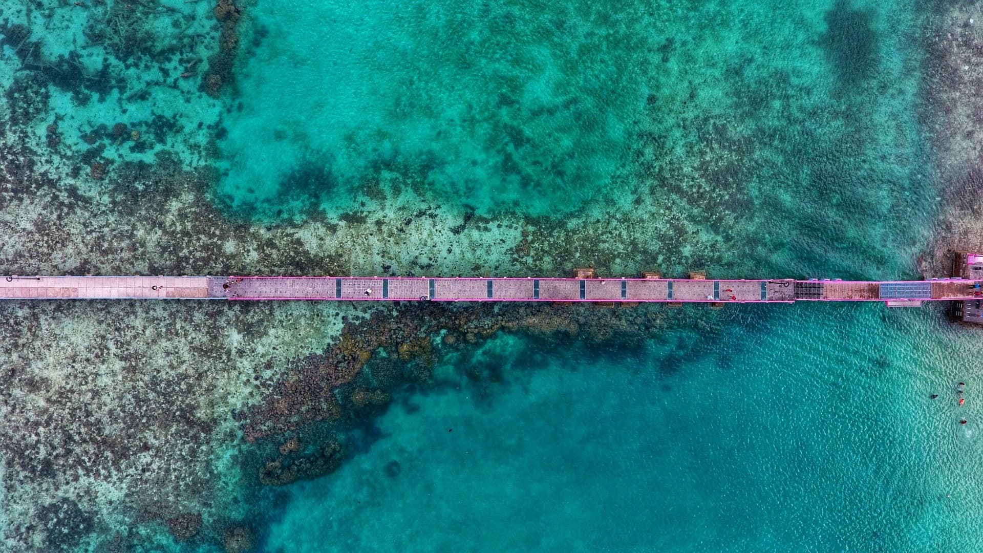 Aerial view of a long pier stretching across shallow turquoise water and dark patches of reef below
