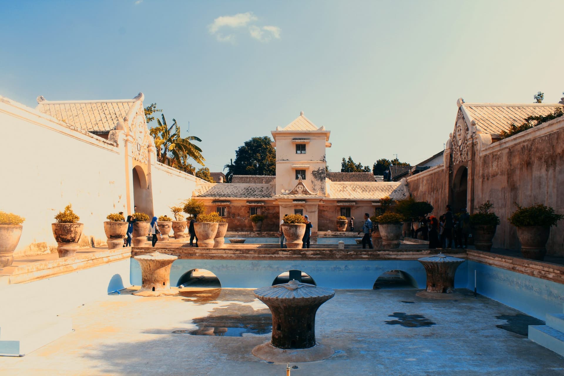 Sunken bathing pool courtyard with round stone platforms, potted plants, and a tiered white tower visitors are walking around