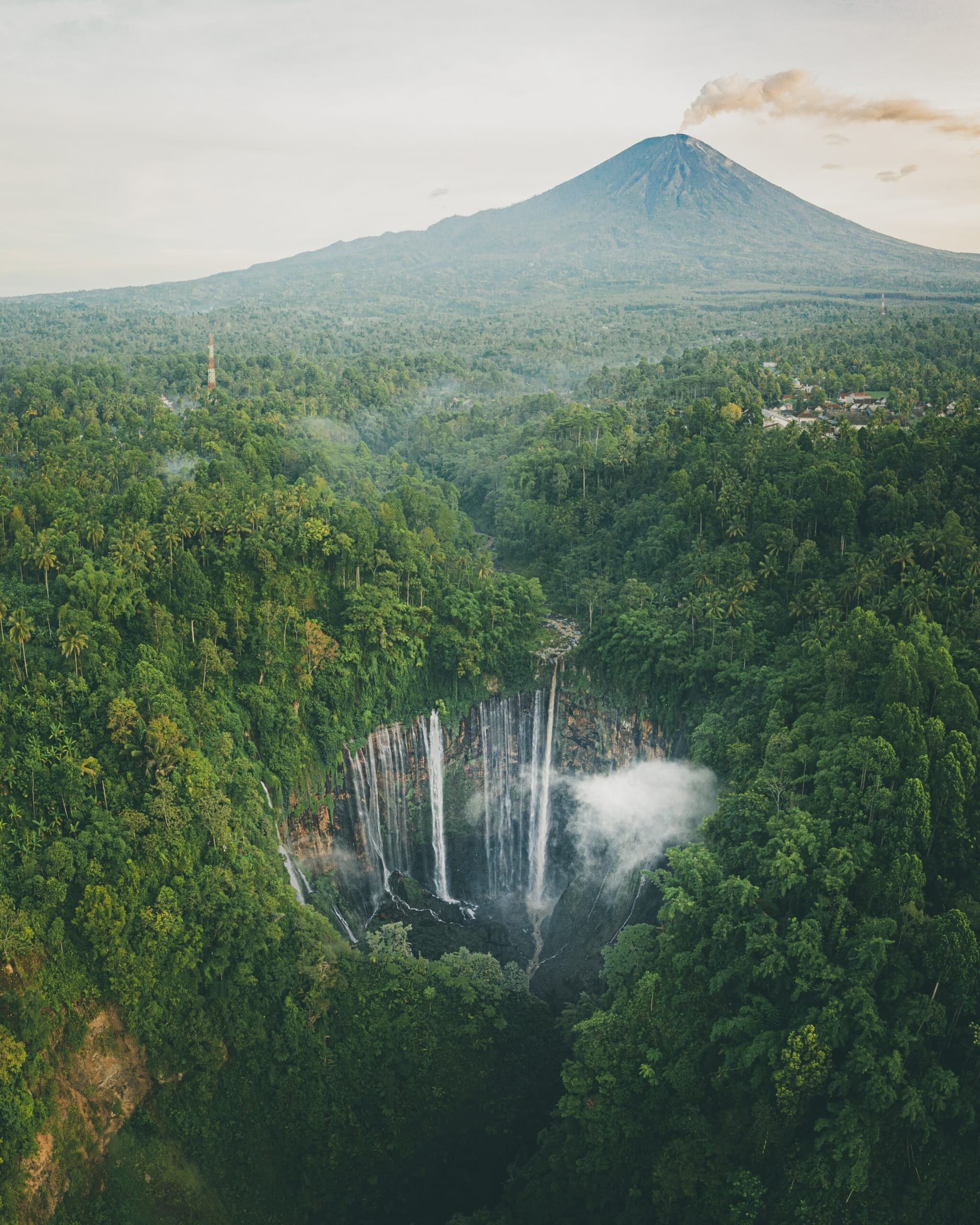 Aerial view of a tall waterfall dropping into a forested gorge with a volcano rising in the distance