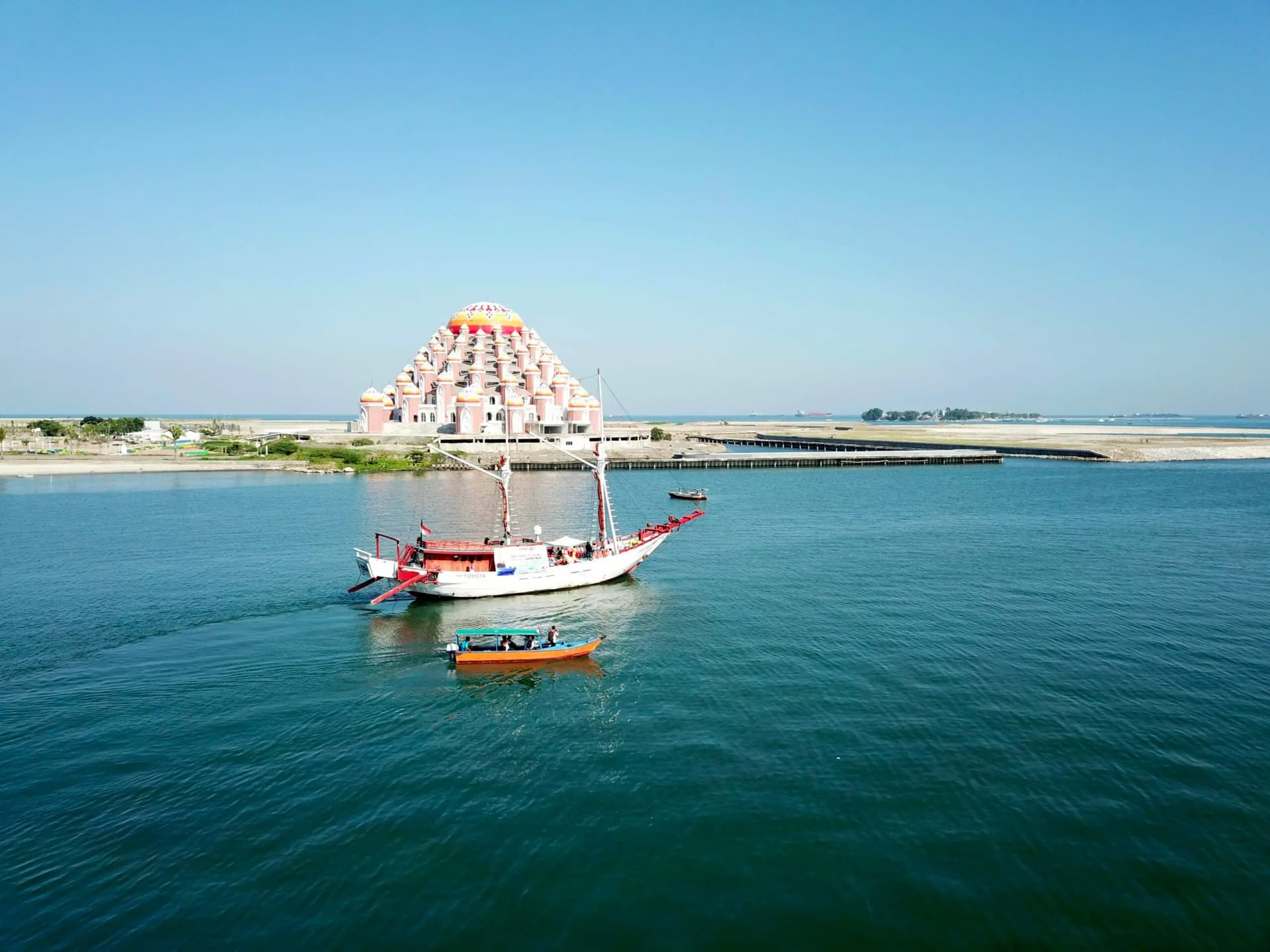 Traditional sailing boat and small motorboat on calm water near a pyramid-shaped building on the shoreline